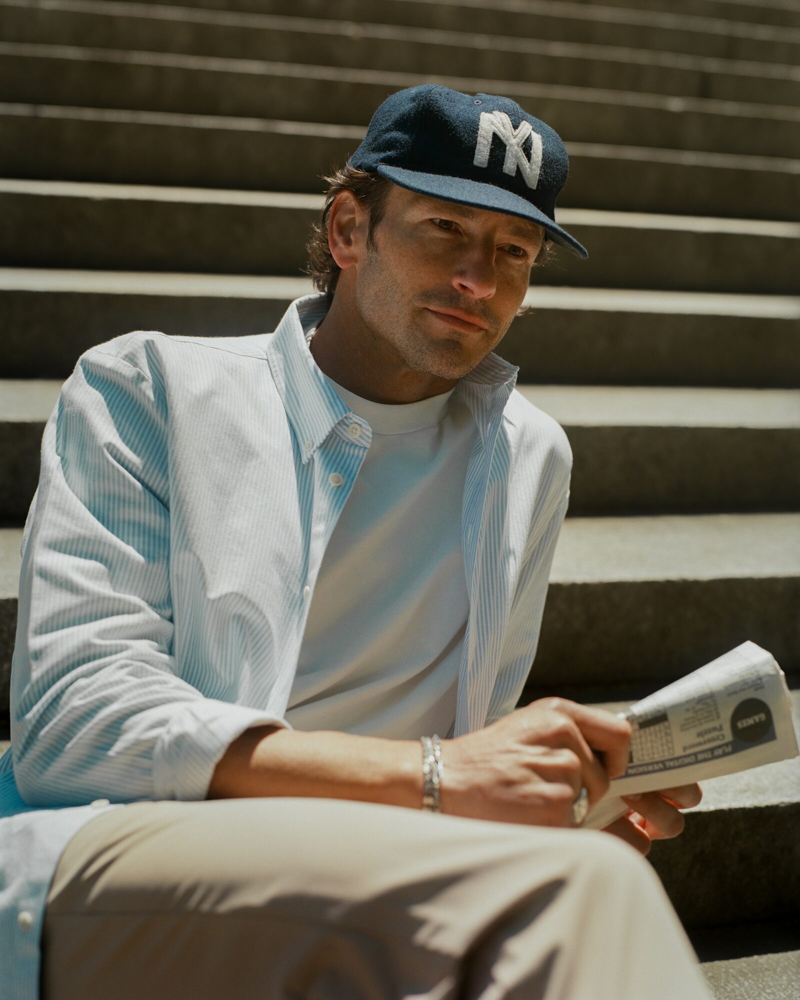 Man sitting on stairs reading a newspaper, wearing a baseball cap and light-colored clothing.