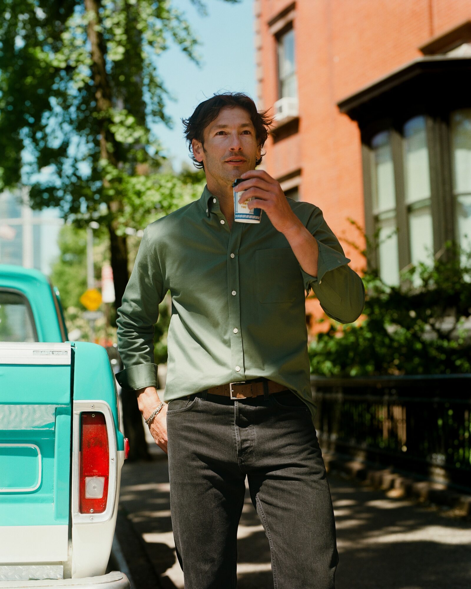 Man standing outdoors on a sunny day, holding a phone near his face, with trees and buildings in the background.