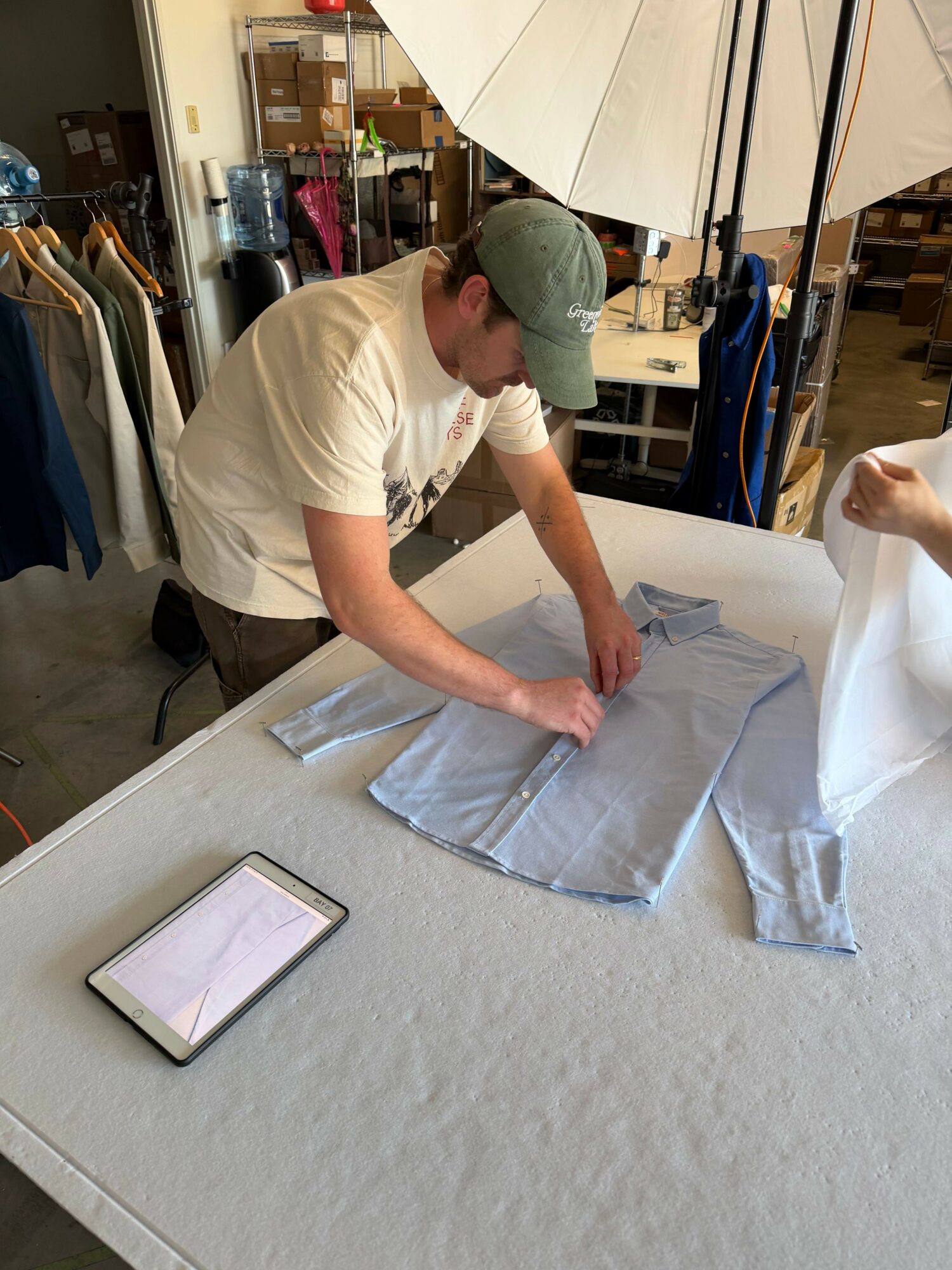 Person folding a light-colored cloth on a table, with a tablet device nearby, in a room with shelves and umbrellas.
