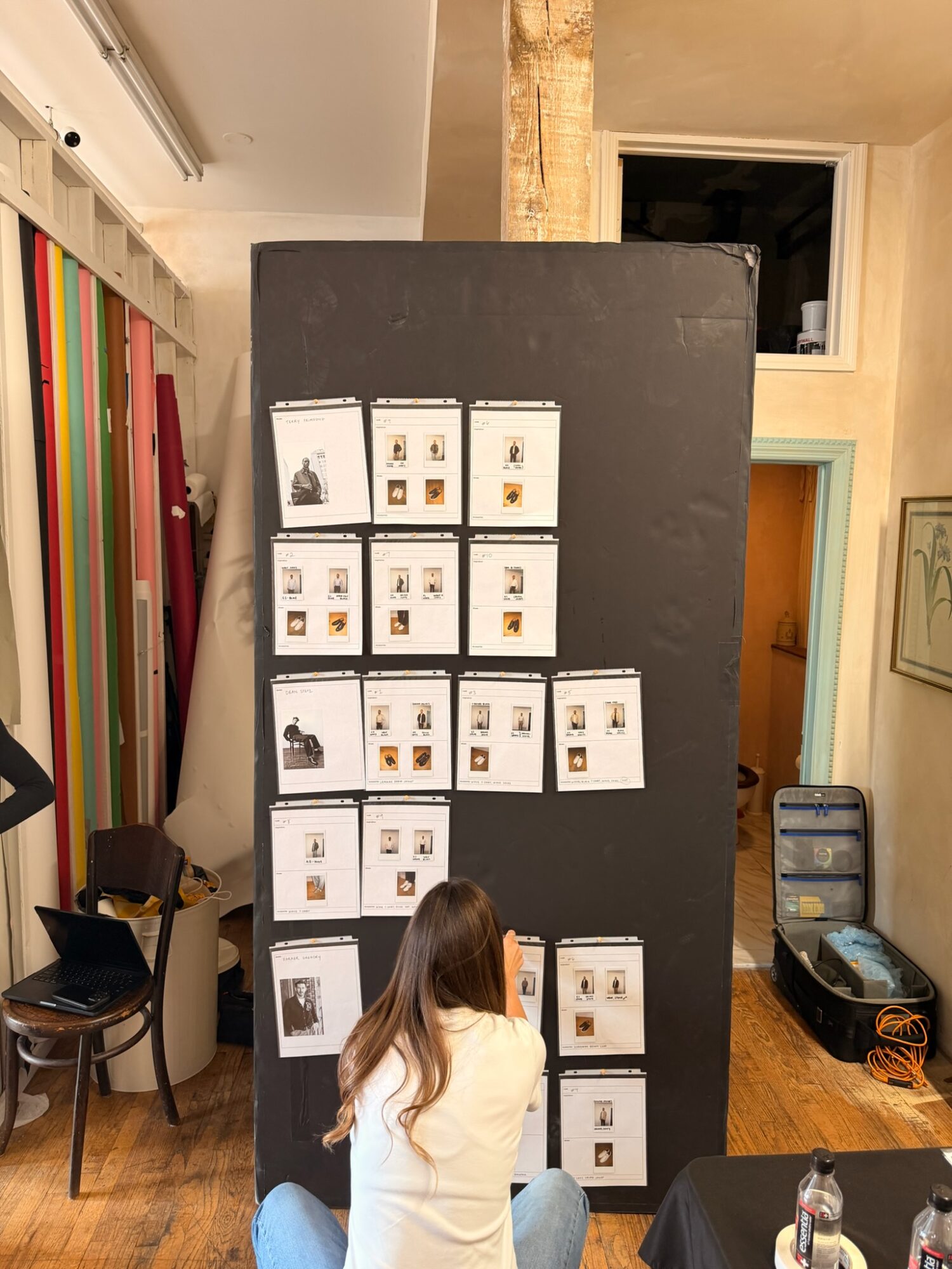 Person with long hair sitting in front of a black display board with printed materials, in a room with colorful vertical stripes on the wall.