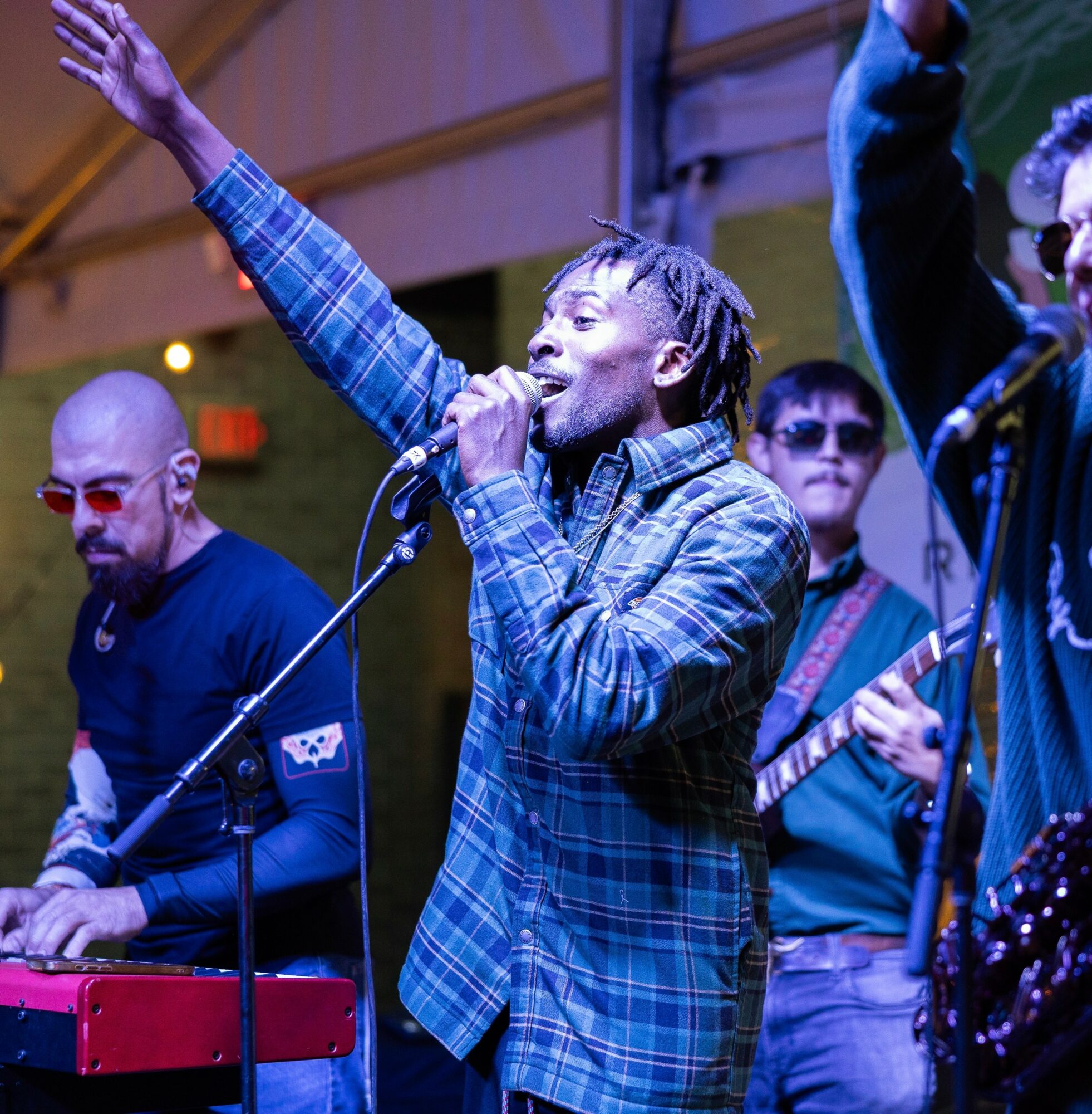 Group of musicians performing on stage, with one singer raising an arm, wearing a plaid shirt, and others playing instruments.