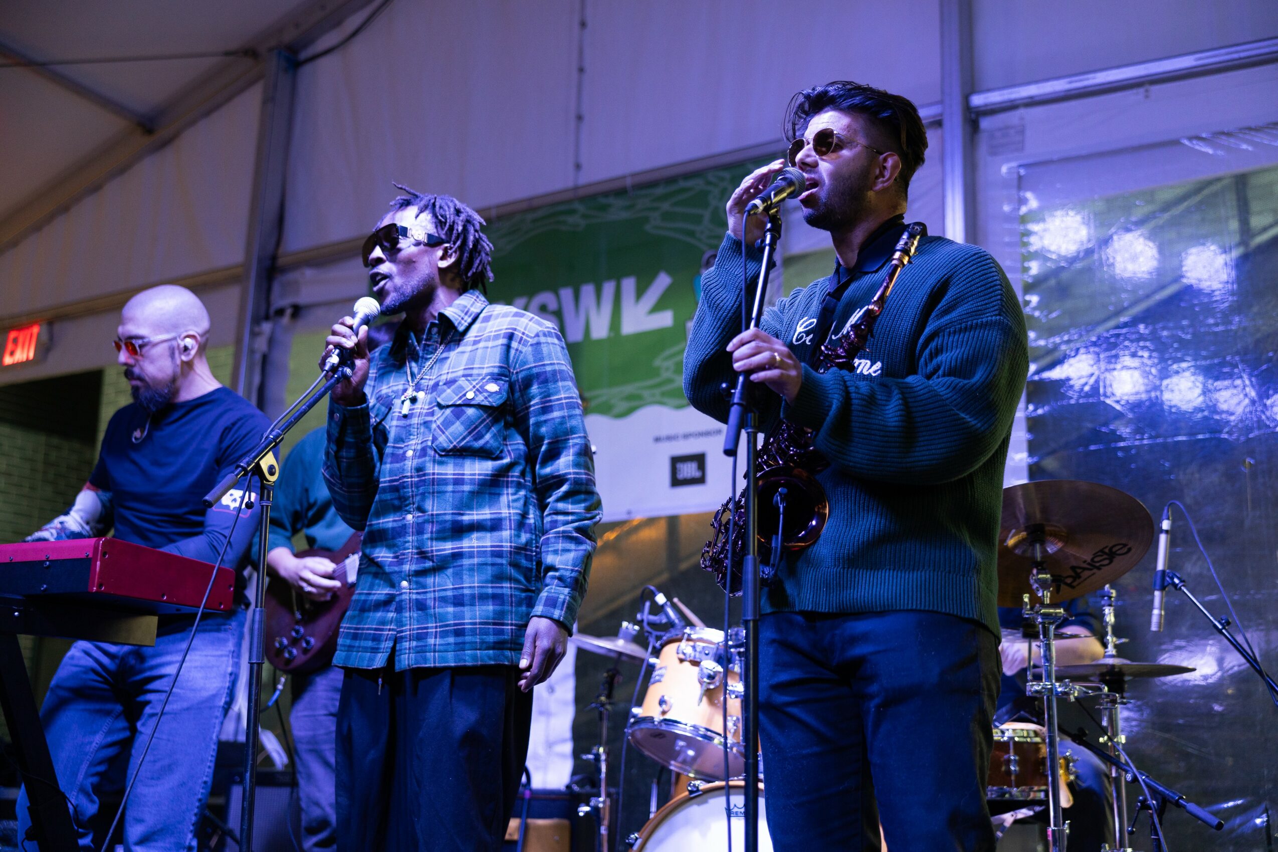Three musicians perform on stage with instruments and microphones, with a drum set in the background, under stage lighting.