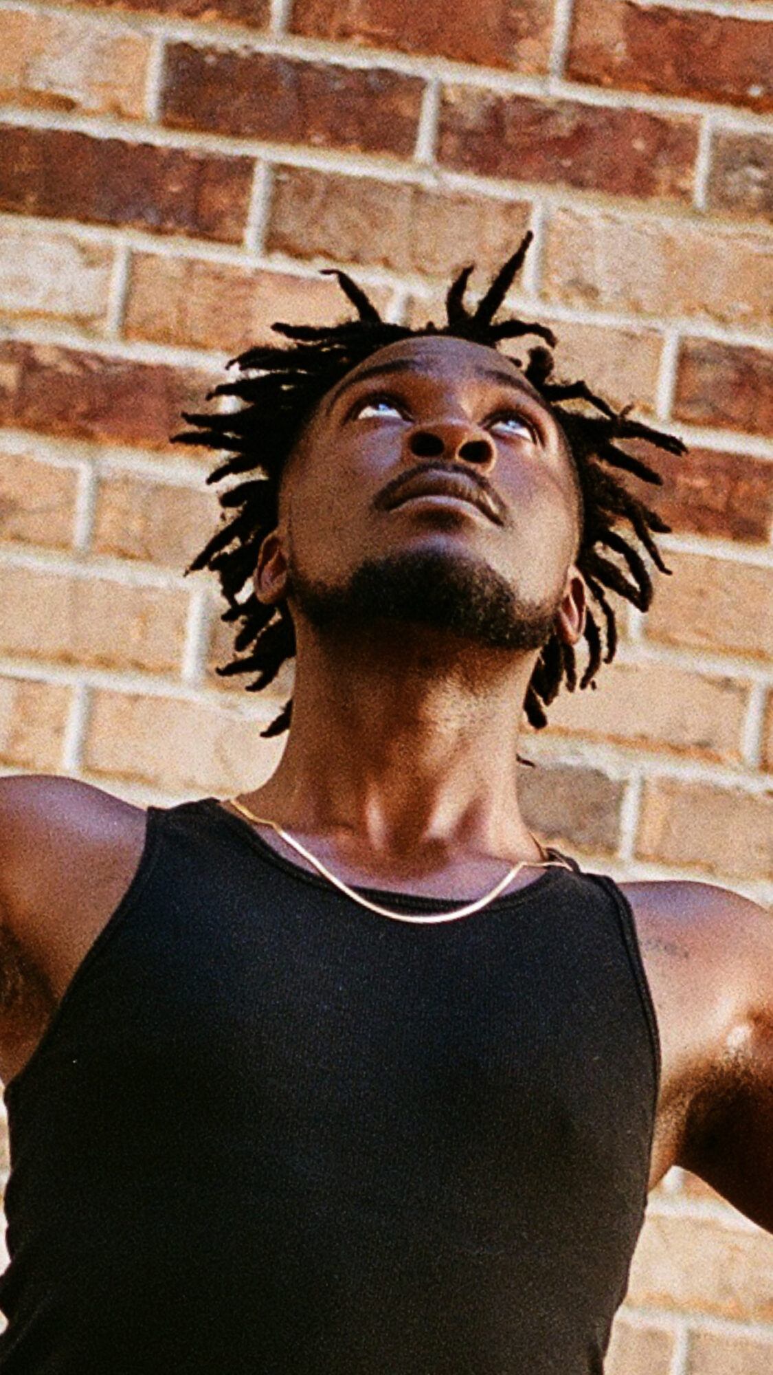 Person with dreadlocks looking upward against a brick wall, wearing a black sleeveless top.