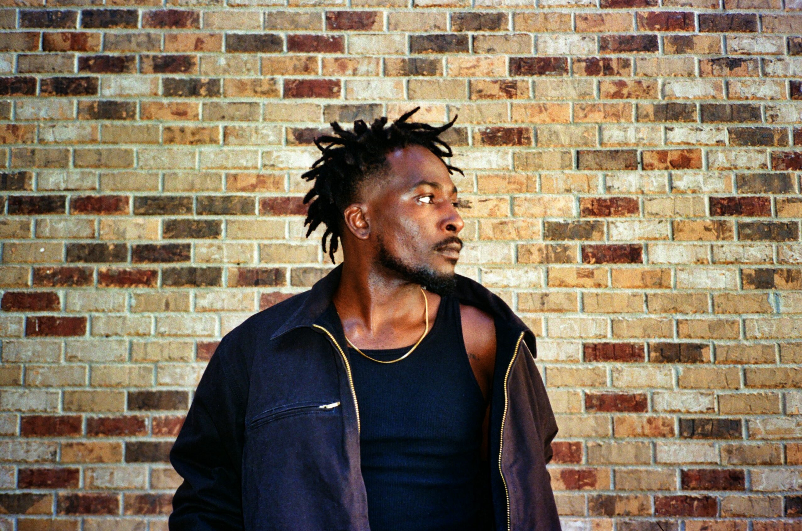 Man with dreadlocks and a beard stands against a brick wall, wearing a black shirt and jacket.
