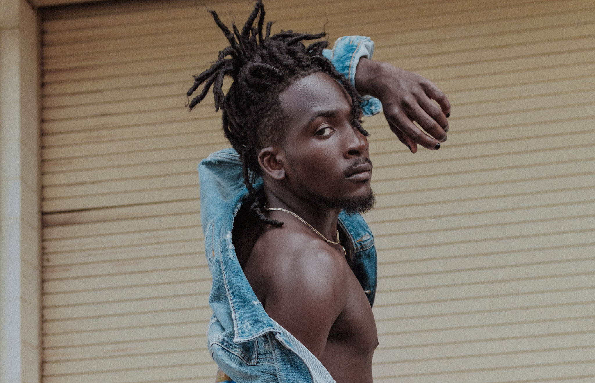 Person with dreadlocks and a denim jacket draped over shoulders, posing against a closed beige shutter.