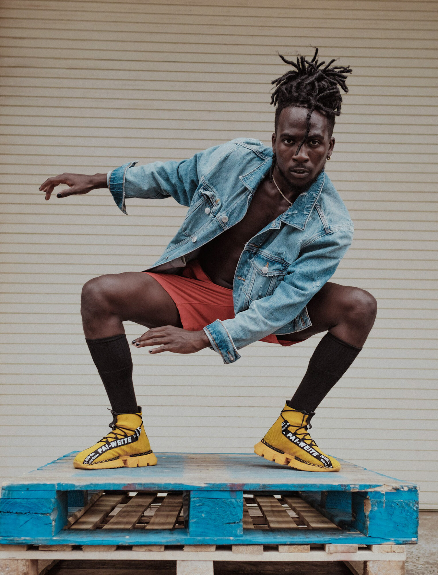 Person crouching on a blue pallet, wearing yellow shoes, red shorts, and a denim jacket, with a neutral background.