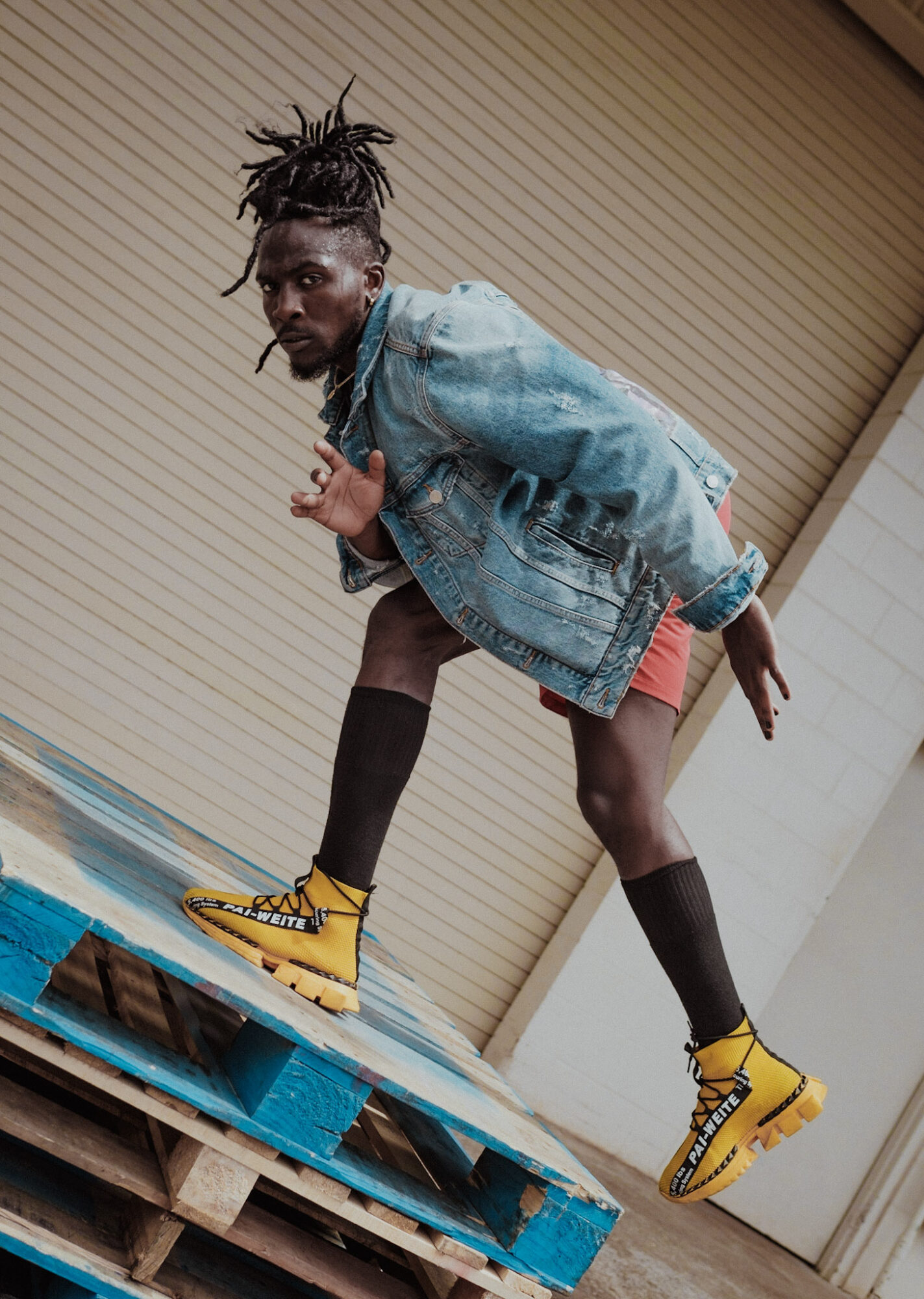 Person with dreadlocks in a denim jacket and yellow boots crouches on a pallet, leaning forward with one hand on knee.