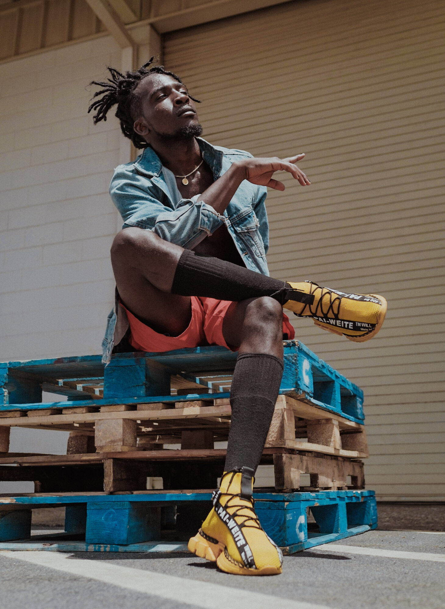 Person with dreadlocks sitting on stacked pallets, wearing colorful clothing and yellow shoes, in an industrial setting.