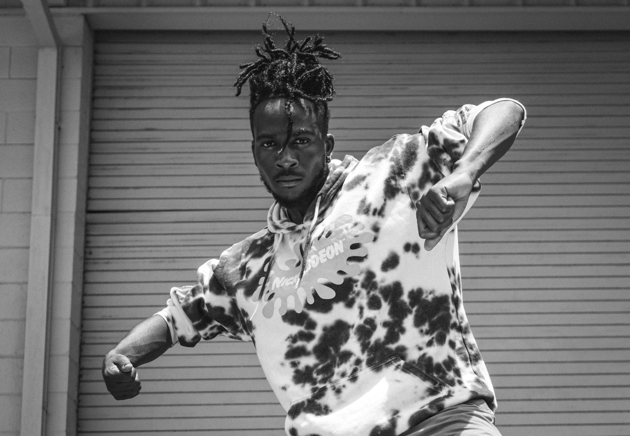 Person with dreadlocks in a tie-dye shirt poses with arms extended in front of a closed garage door.