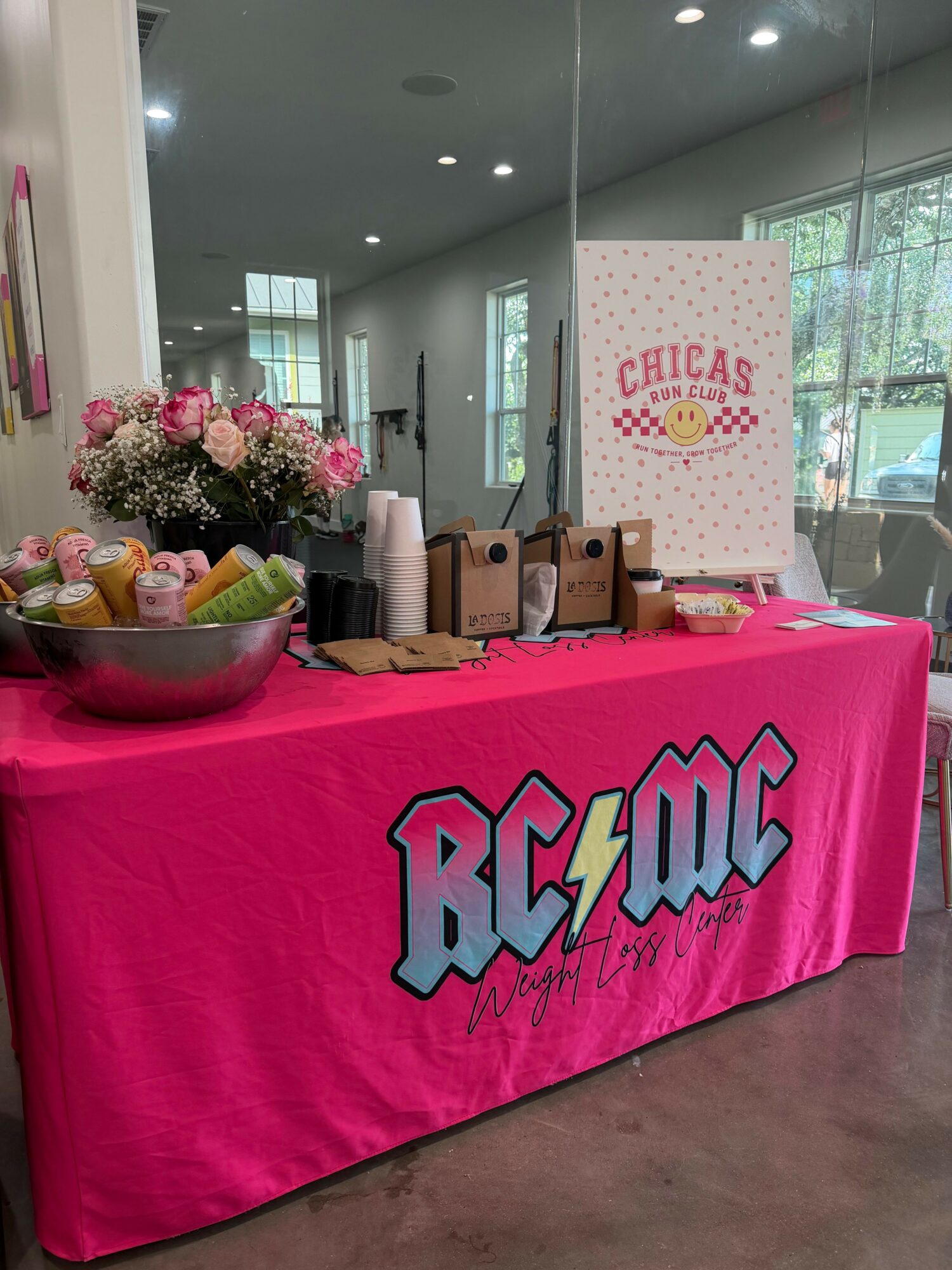 Table with pink tablecloth featuring the word 'BOOM' in large letters, decorated with a flower arrangement and party supplies, inside a modern room.