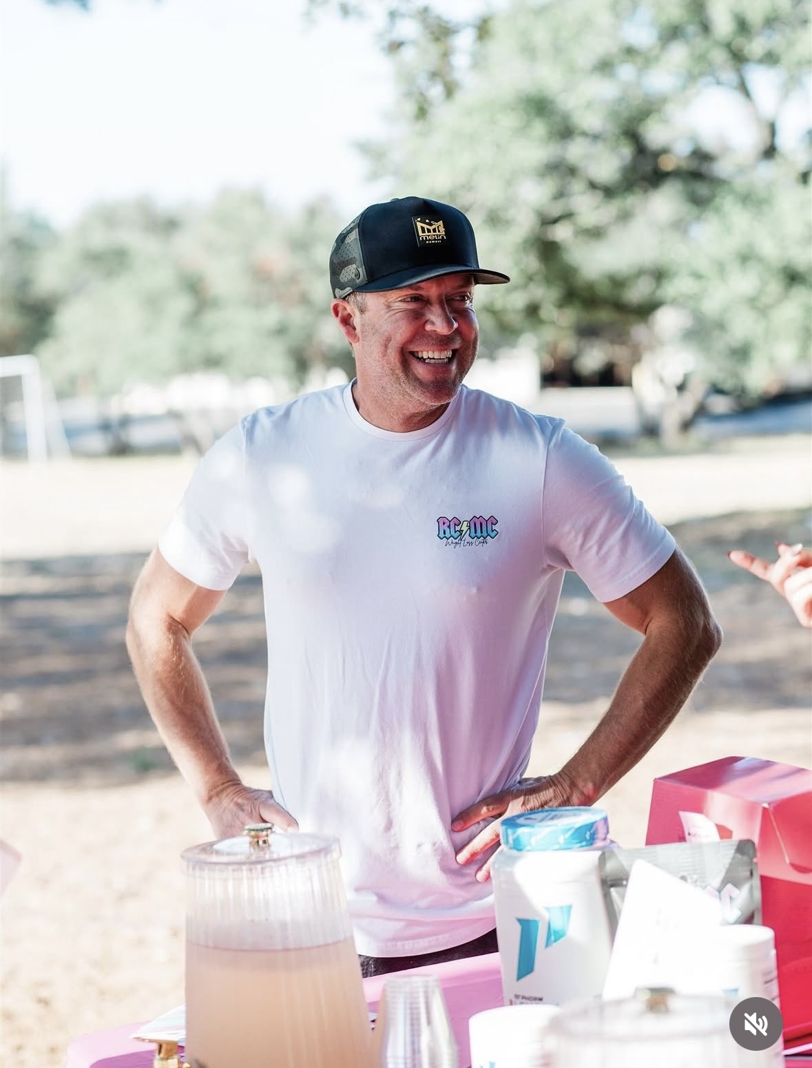 Person smiling outdoors, wearing a white t-shirt and a black cap, standing near a table with drinks.