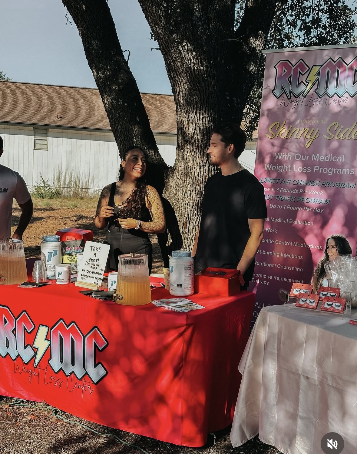 Two people standing behind a red table with a large tree in the background, outdoor event, sunny day.