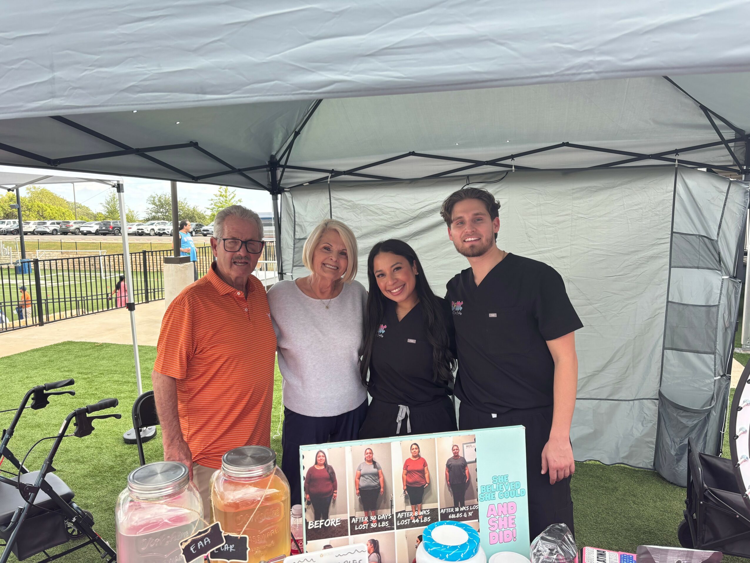 Four people standing inside a tent, smiling, with a table displaying photos and drinks, outdoors on a grassy field.