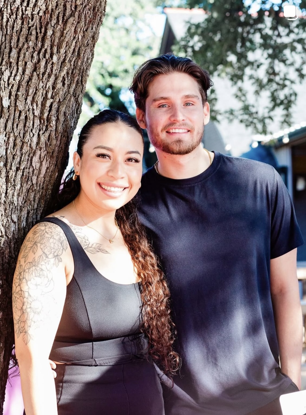 A smiling woman with long curly hair and a man with short hair and beard stand outdoors near a tree.
