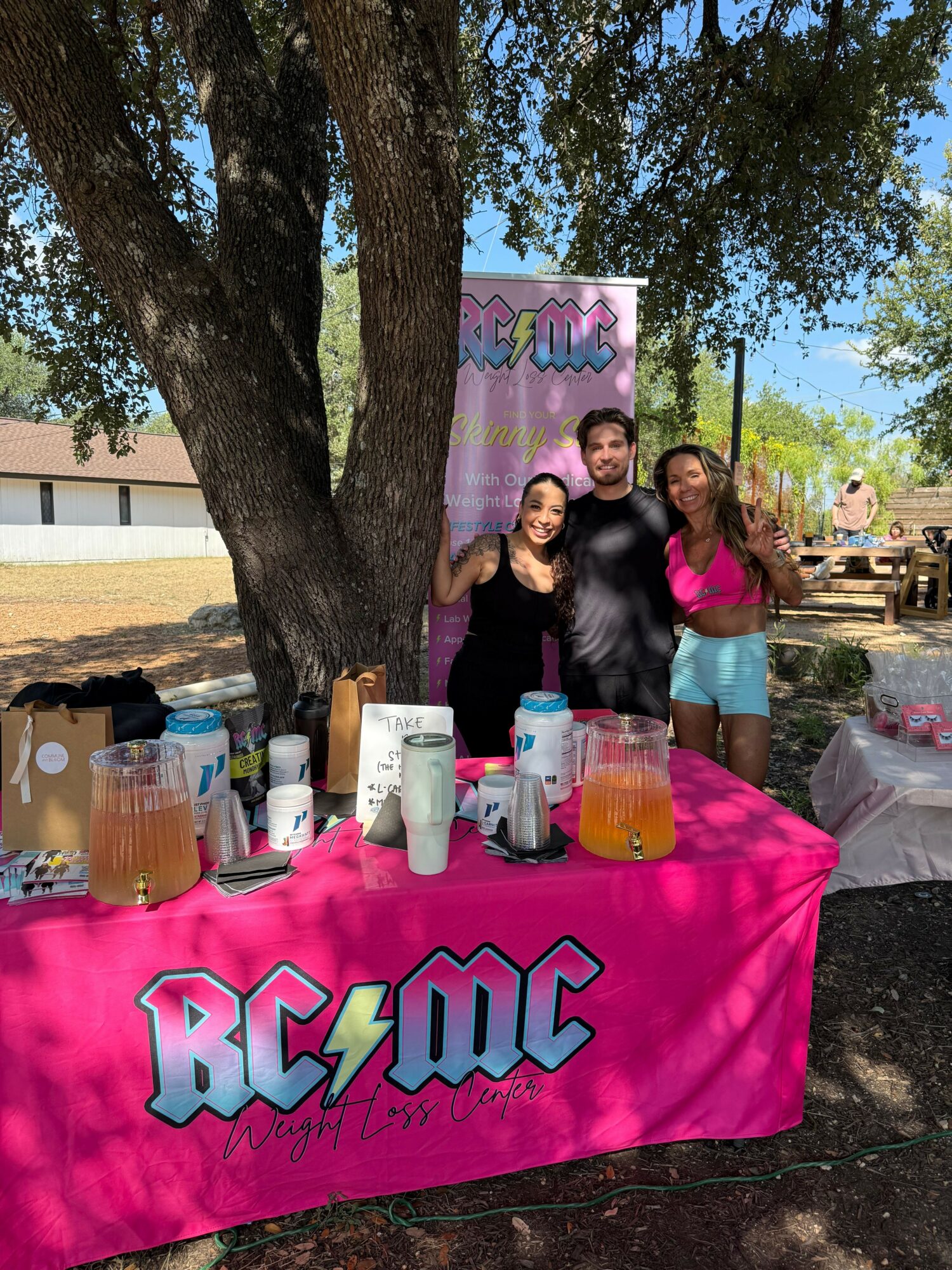 Three women stand behind a table with drinks and a pink tablecloth that says 'BECAUSE'. They are outdoors under a tree.