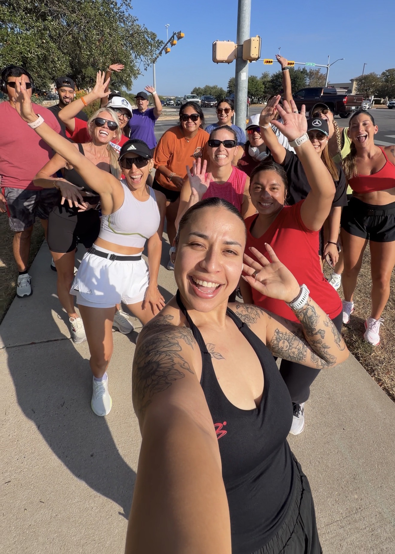Group of smiling women posing outdoors on a sunny day, some making peace signs, with trees and a street in background.