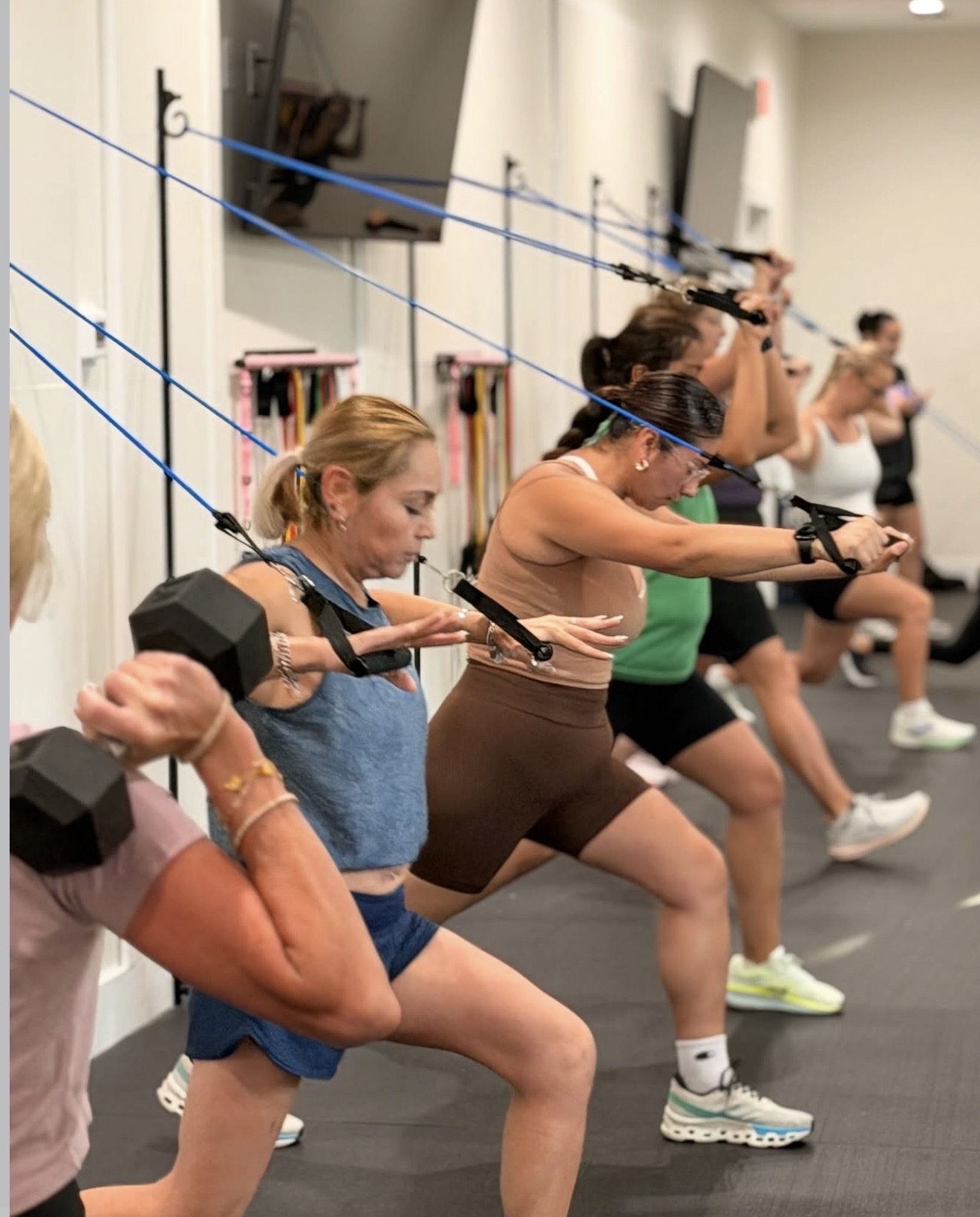 Group of women exercising with resistance bands attached to the wall, performing leg stretches in a fitness class.