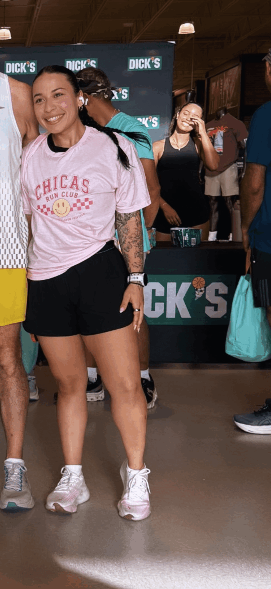 Young woman smiling, wearing a pink T-shirt and black shorts, standing indoors with people and a Dick's sign in background.