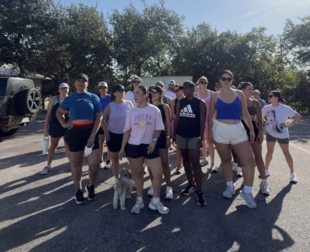 Group of people standing outdoors in a parking lot with trees in the background, some wearing athletic clothing.