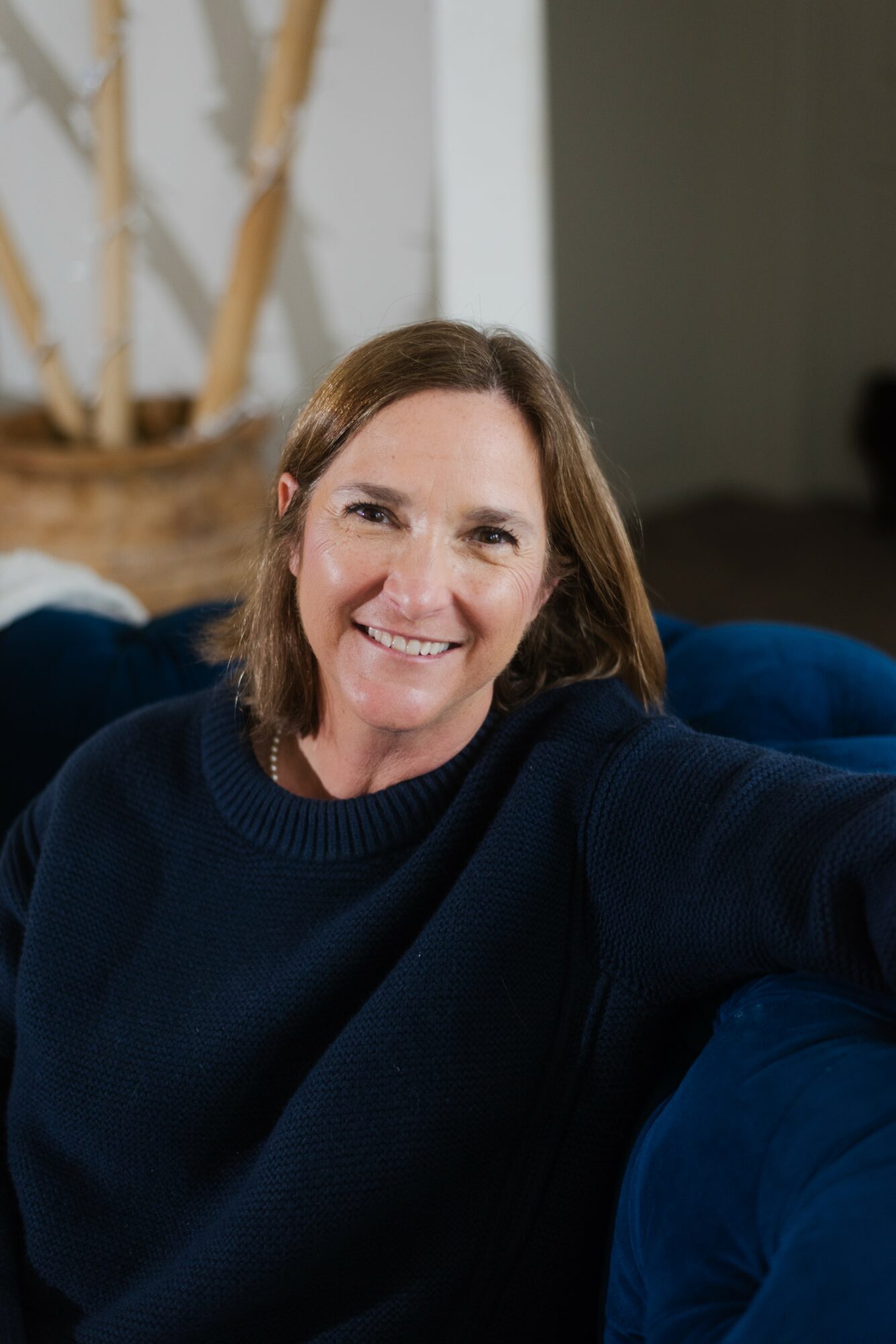Woman with shoulder-length brown hair smiling, wearing a dark sweater, sitting on a blue couch indoors.