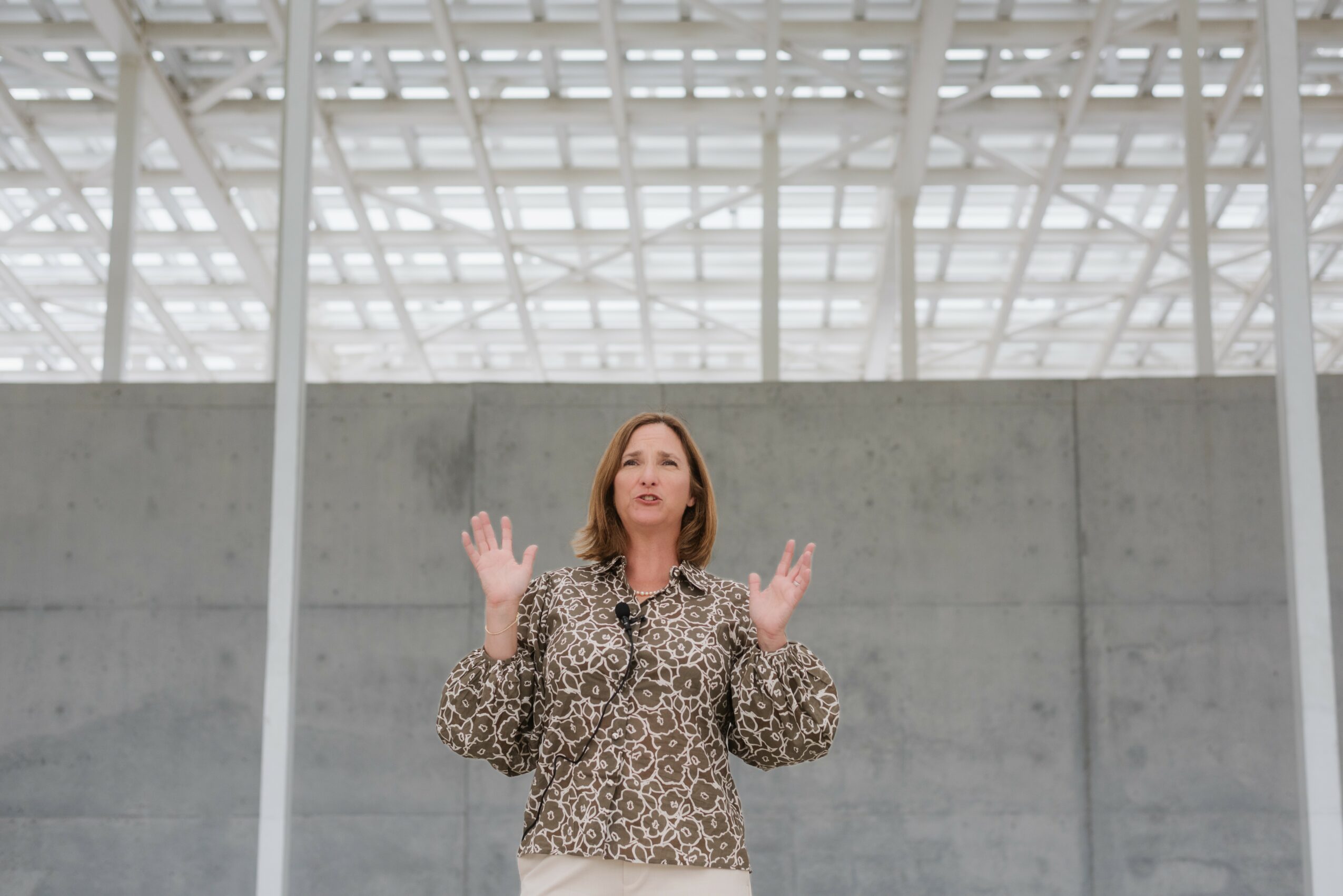 Woman standing indoors with arms raised, wearing a leopard print blouse, in a modern building with glass ceiling and concrete wall.