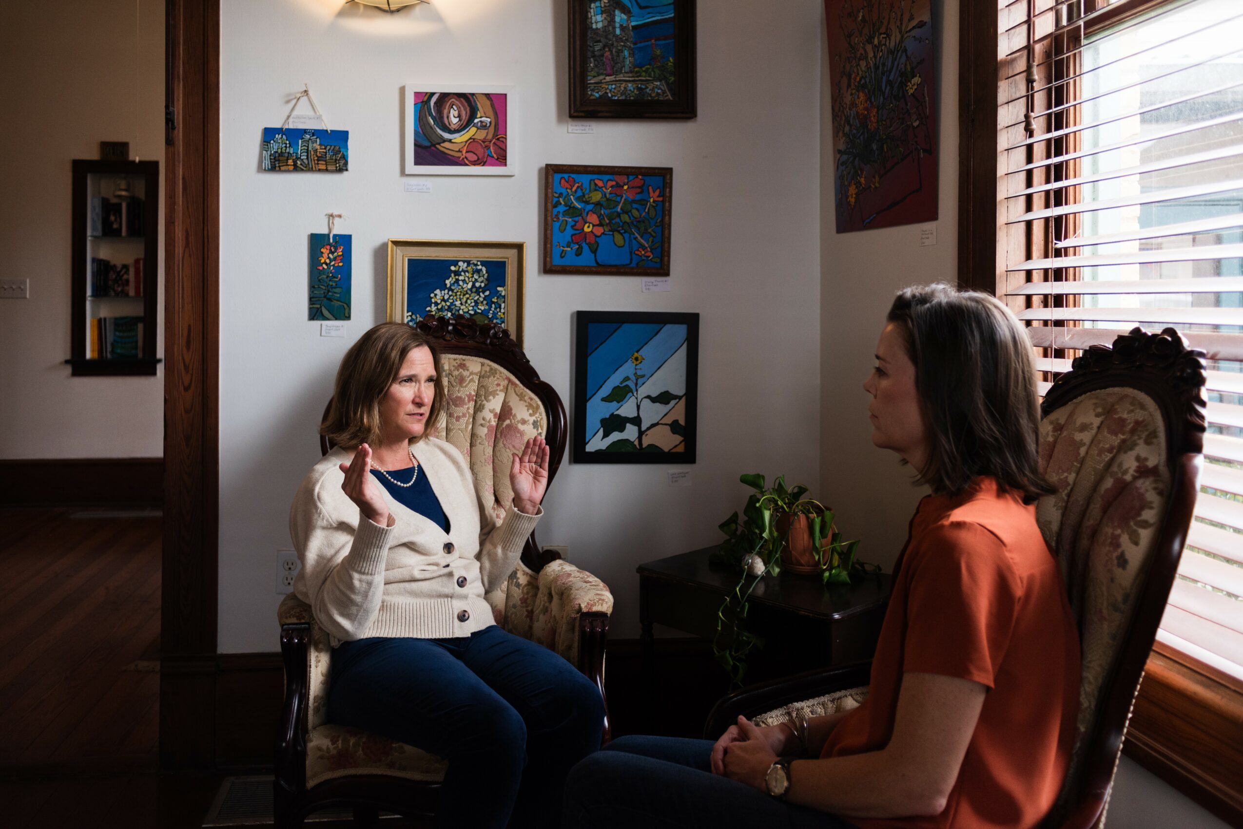 Two women sit in chairs facing each other in a room with artwork on the wall and a window with blinds.