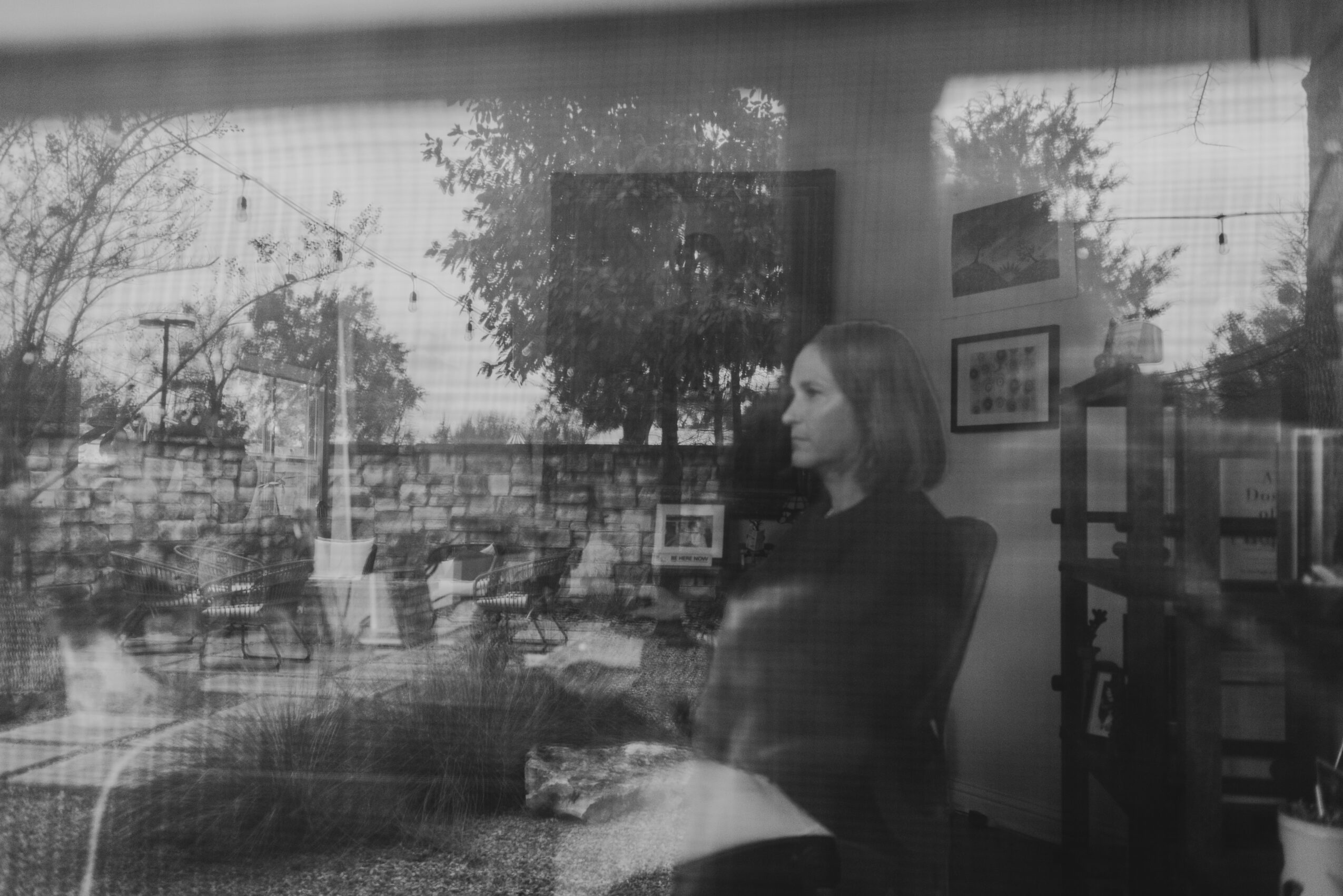 A woman sitting indoors near a window with trees and outdoor scenery visible, holding a book or object in her lap.