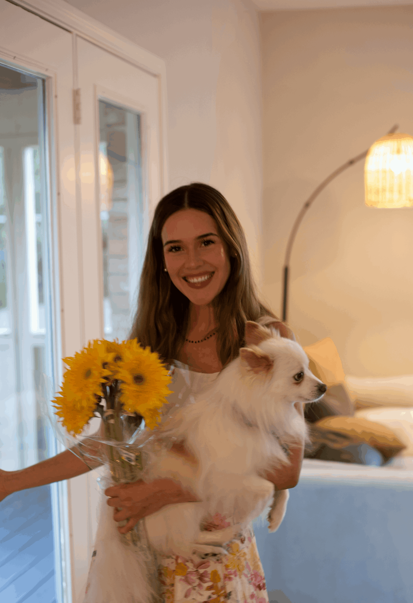 Smiling woman holding a small dog indoors, with a person offering a bouquet of yellow flowers nearby.