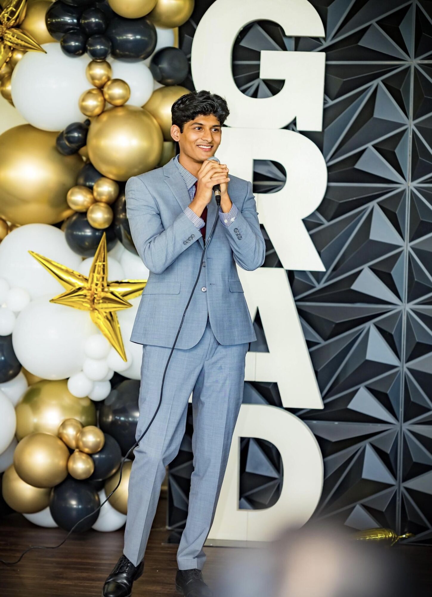 Young man in a light blue suit speaking into a microphone at a decorated event with balloons and geometric backdrop.