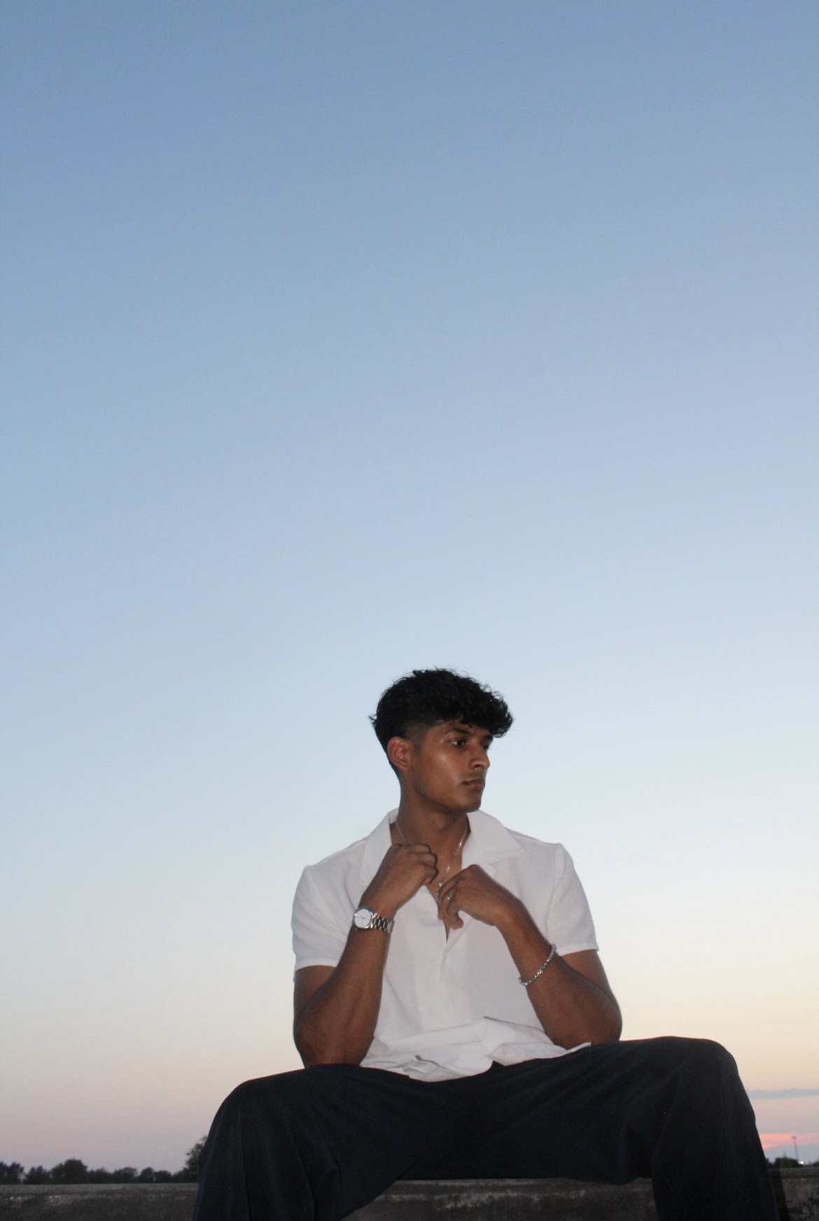 Young man sitting outdoors against a clear sky, wearing a white shirt and black pants, looking to the side.