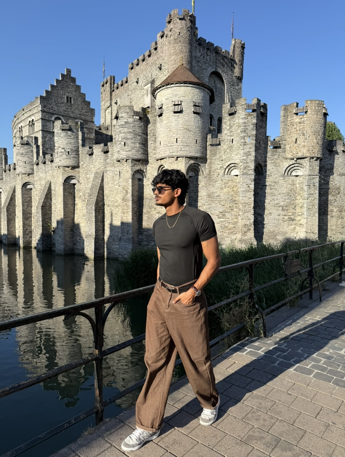 Young man in sunglasses and casual clothing stands by a river with a historic castle in the background.