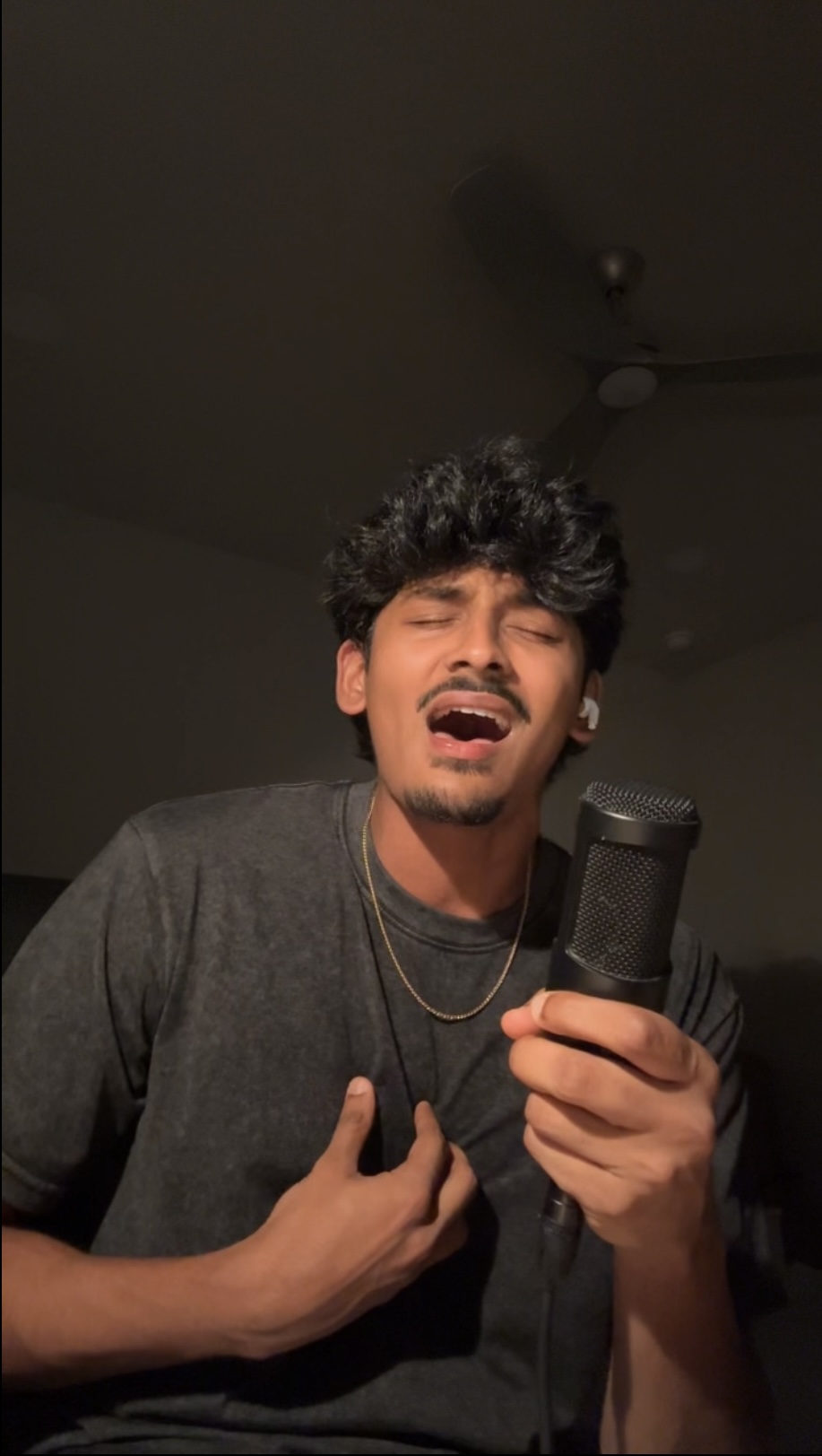 Young man singing into a microphone, eyes closed, hand on chest, in a dimly lit room.