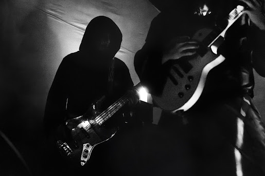Two people playing electric guitars on stage, one in shadow, with stage lighting creating silhouettes.