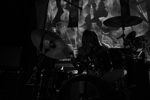 Musician playing drums on stage with a projected image in the background, black and white, dim lighting