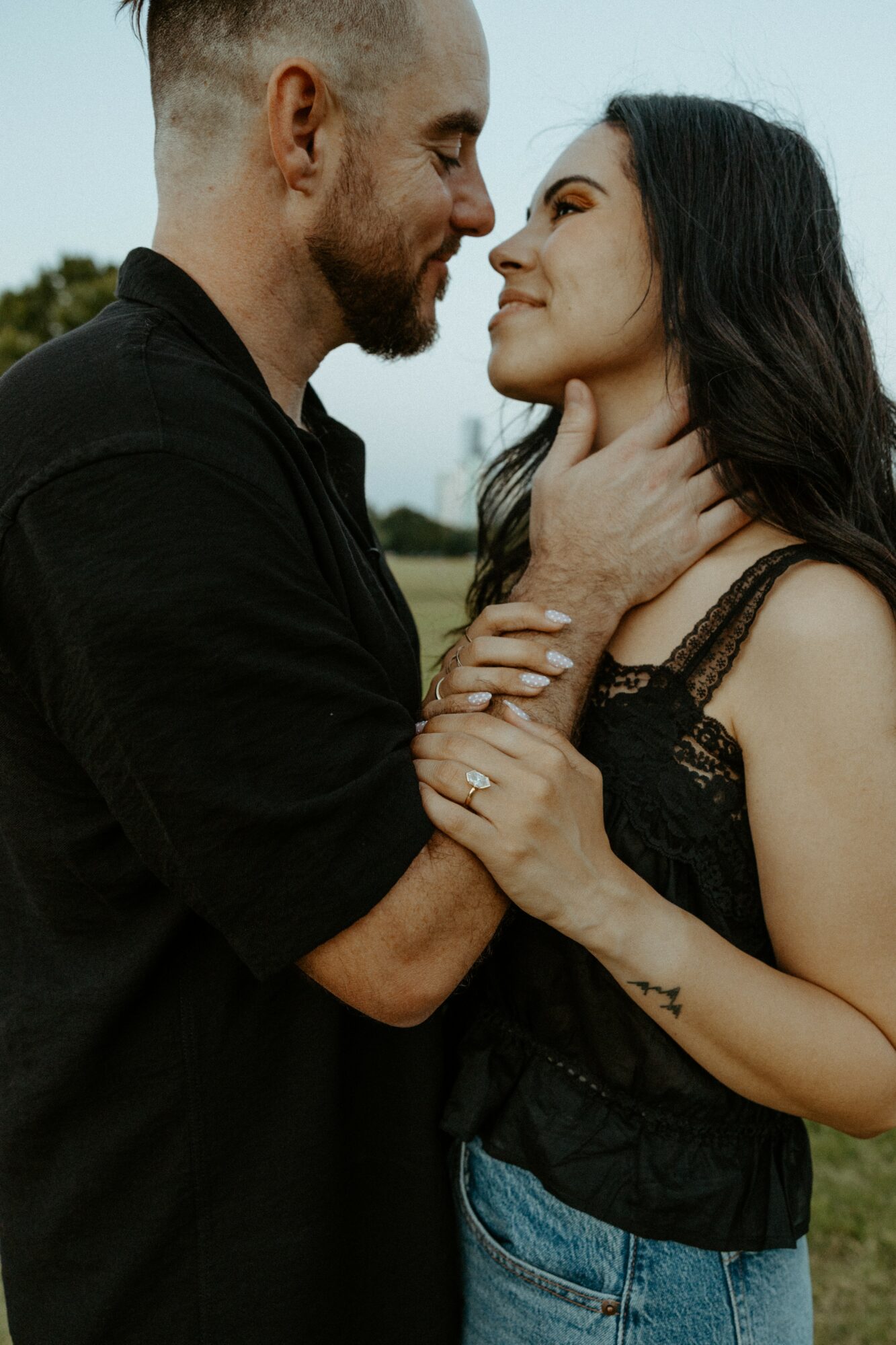 A man and woman face each other closely, holding hands, outdoors with a cloudy sky background.