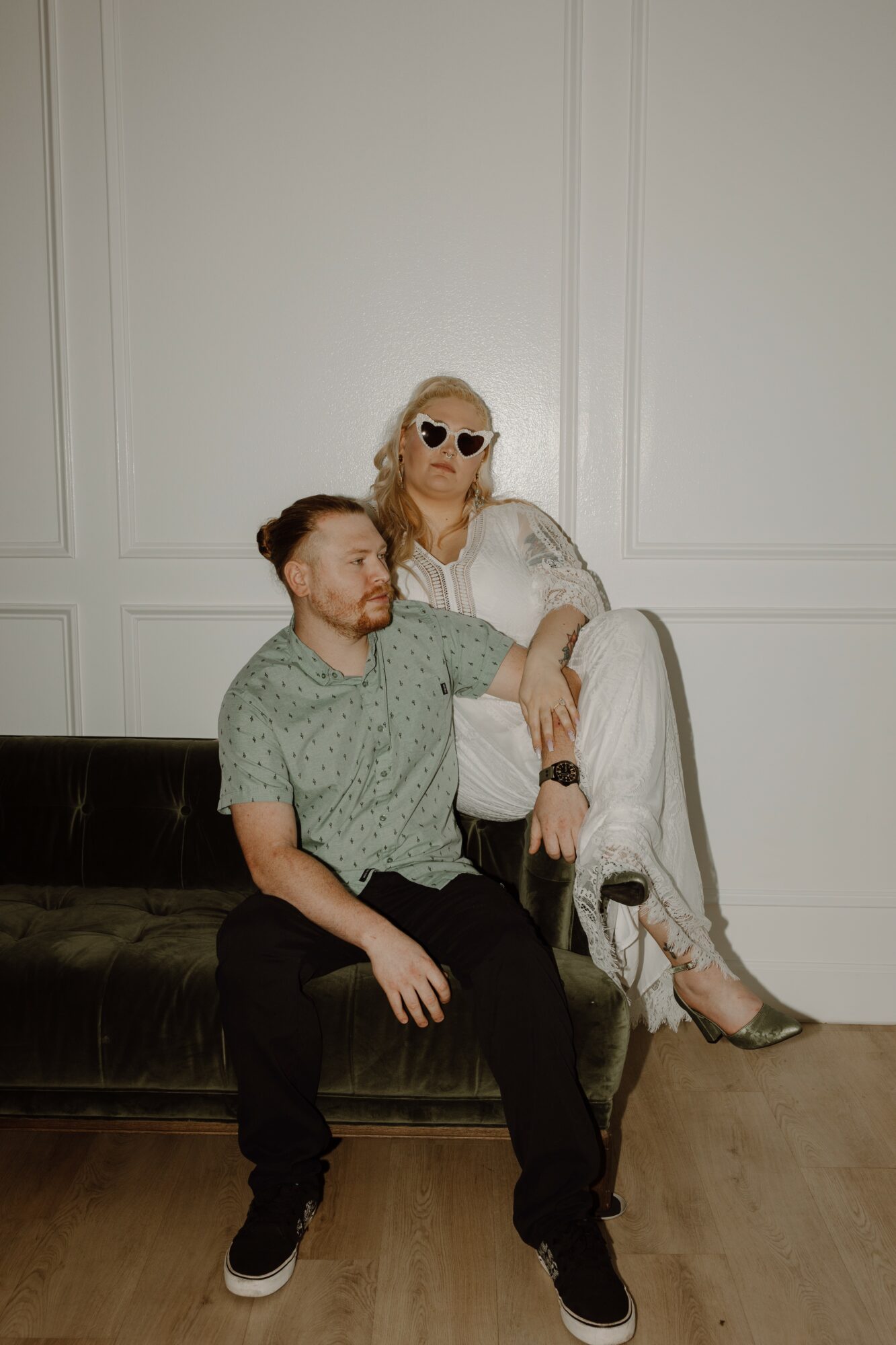 A man and woman sit on a black sofa against a white wall, with the woman wearing sunglasses and white clothing.