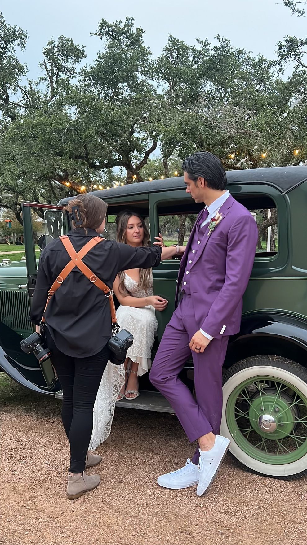 Three people near a vintage green car, one in a purple suit, two in casual clothing, outdoors with trees.