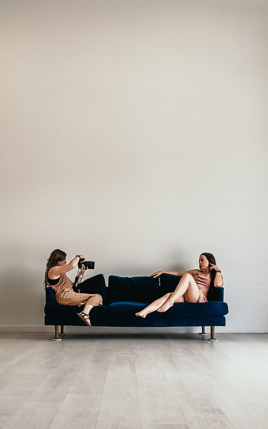 Two women on a dark blue sofa, one taking a photo of the other, against a plain white wall.