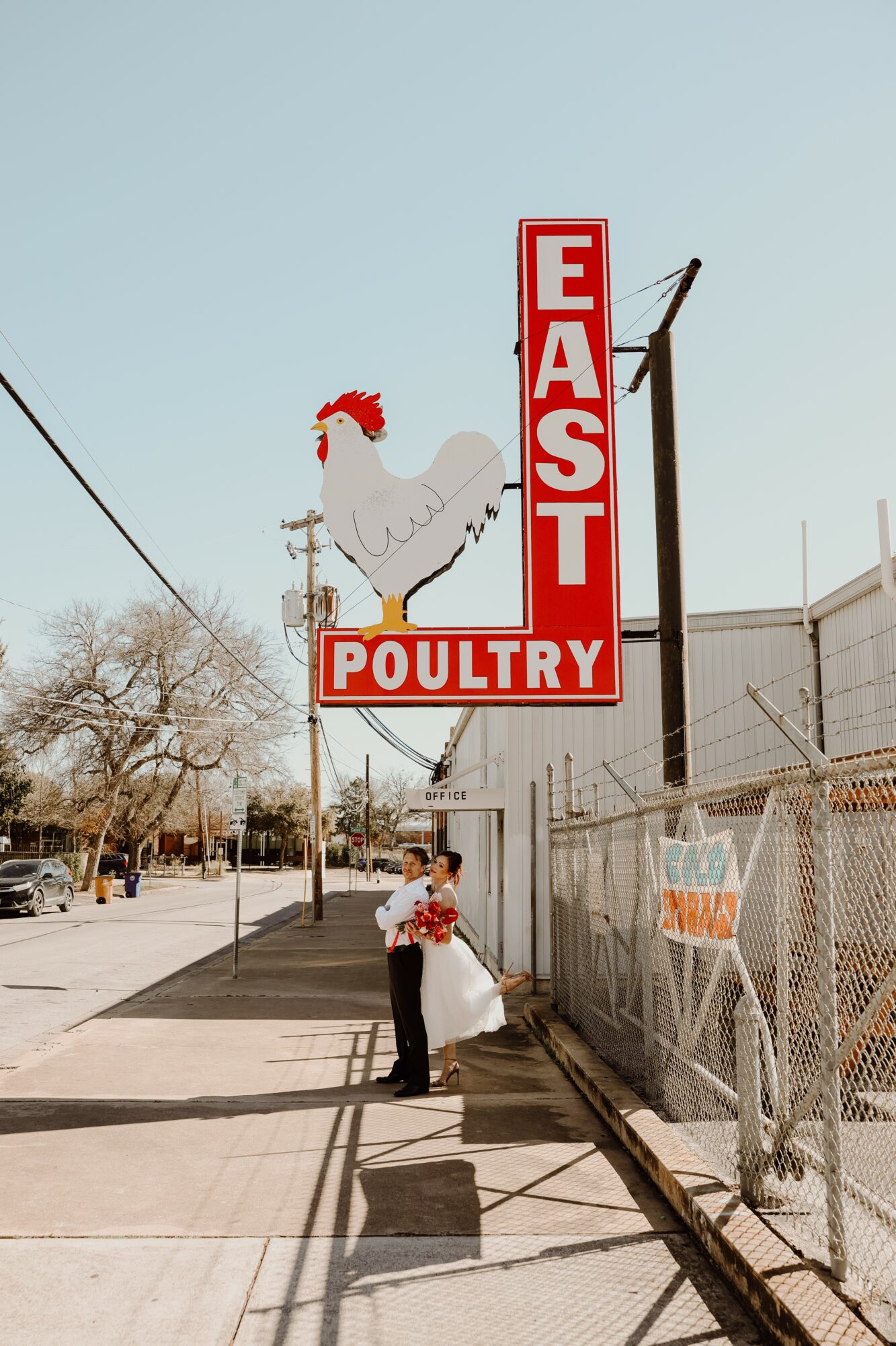 Person holding a bouquet of flowers standing on sidewalk near a poultry sign with a chicken illustration, fence, and trees in background.