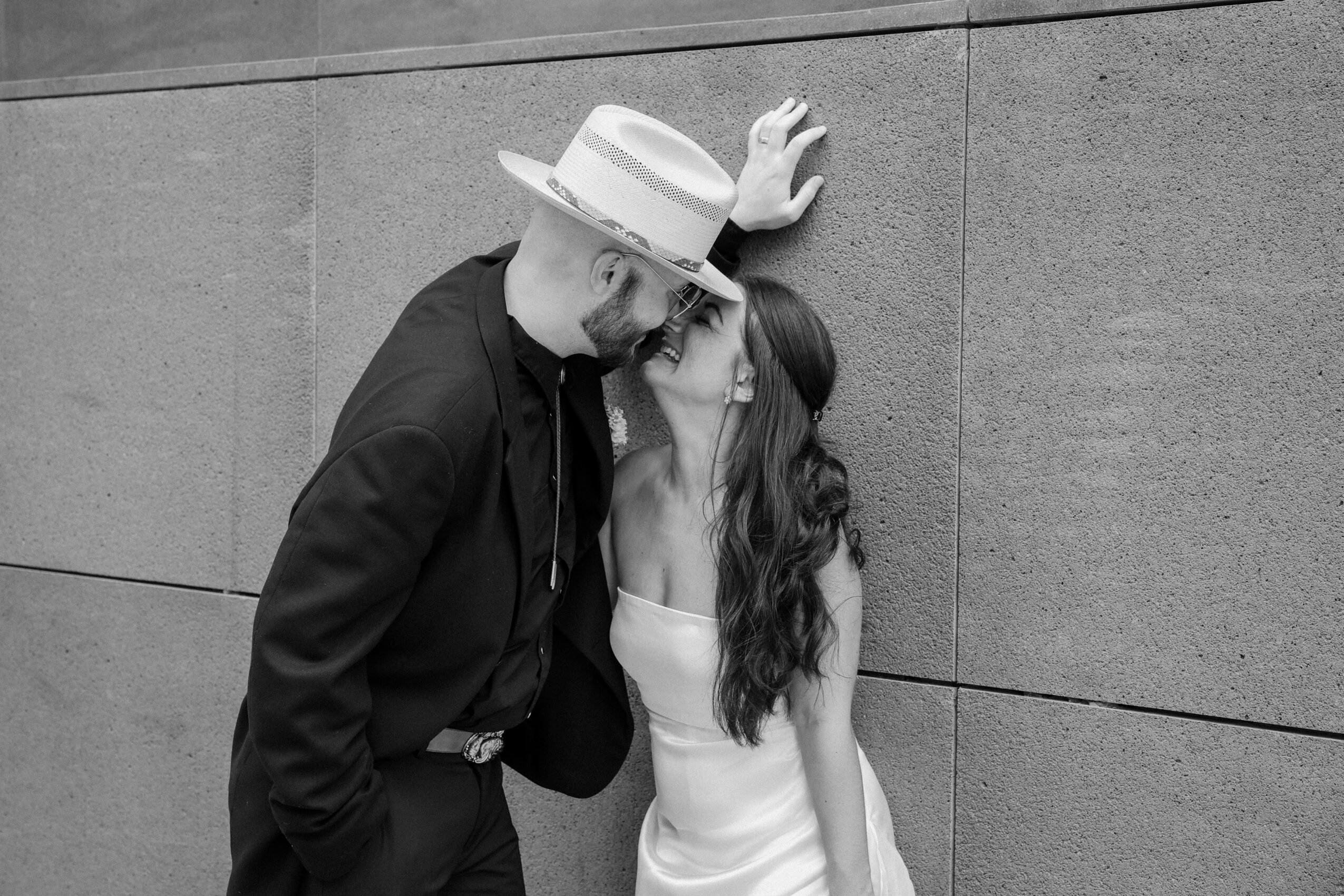 Man and woman leaning against a wall, touching foreheads, man wearing a hat and dark jacket, woman in a white dress.