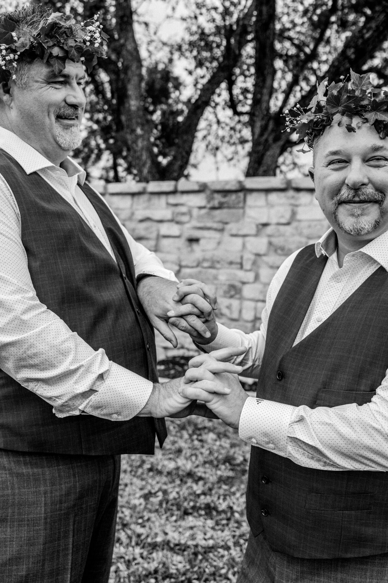 Two men in suits holding hands and smiling outdoors, with a stone wall and trees in the background.