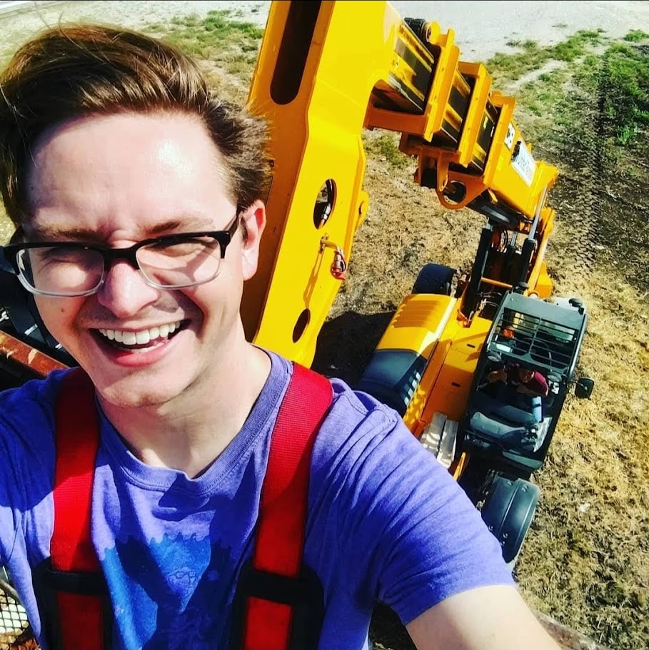 Person smiling with glasses, taking a selfie with construction equipment and a vehicle in the background outdoors.