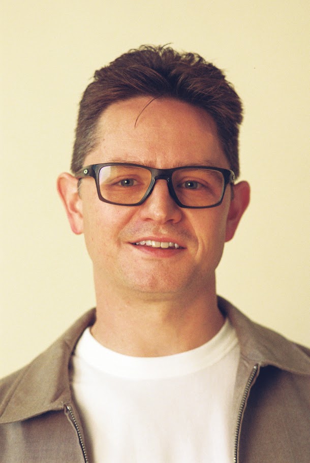 Portrait of a man with glasses, short dark hair, smiling, wearing a white shirt and gray jacket, against a light background.