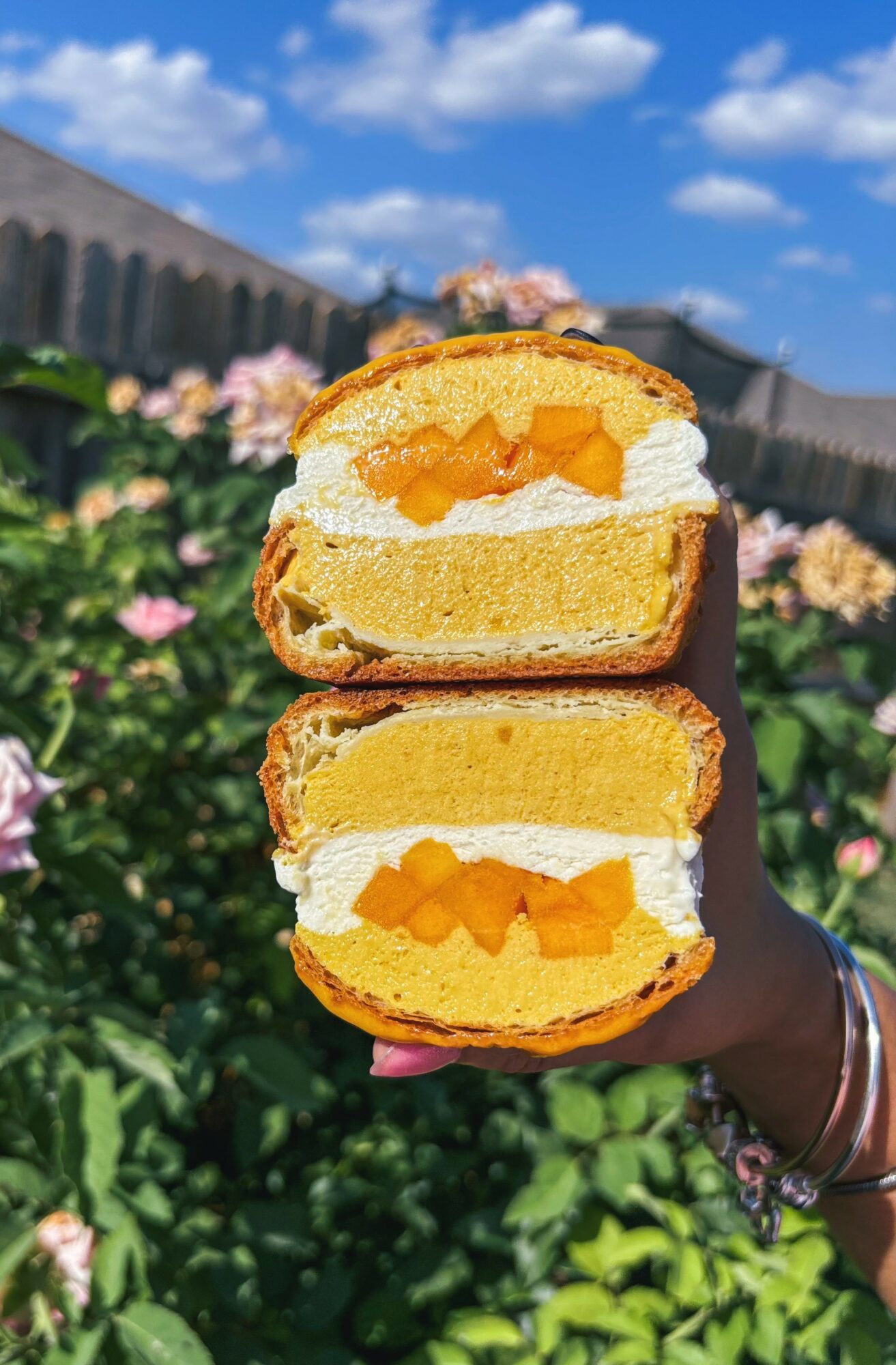 Hand holding a sliced pastry with cream and fruit filling against a garden background with flowers and blue sky.
