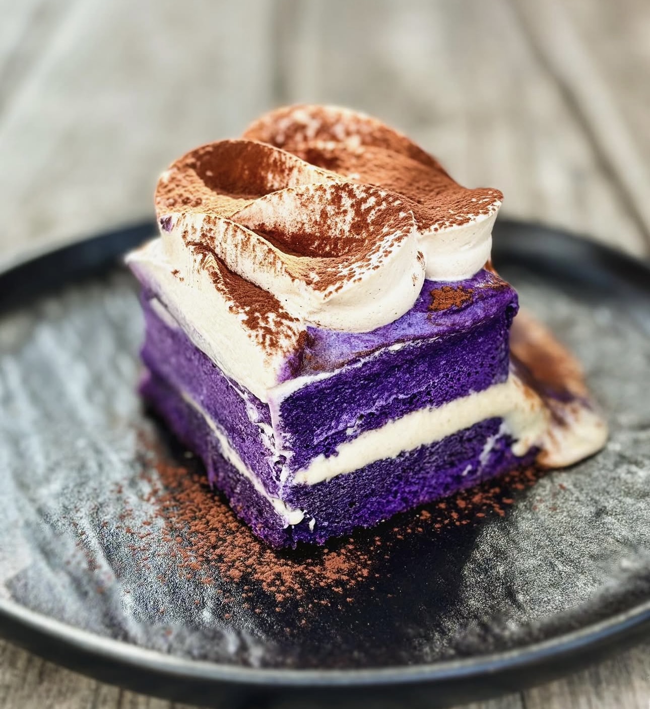 A layered dessert with purple, white, and cream-colored layers topped with whipped cream and cocoa powder, served on a black plate.