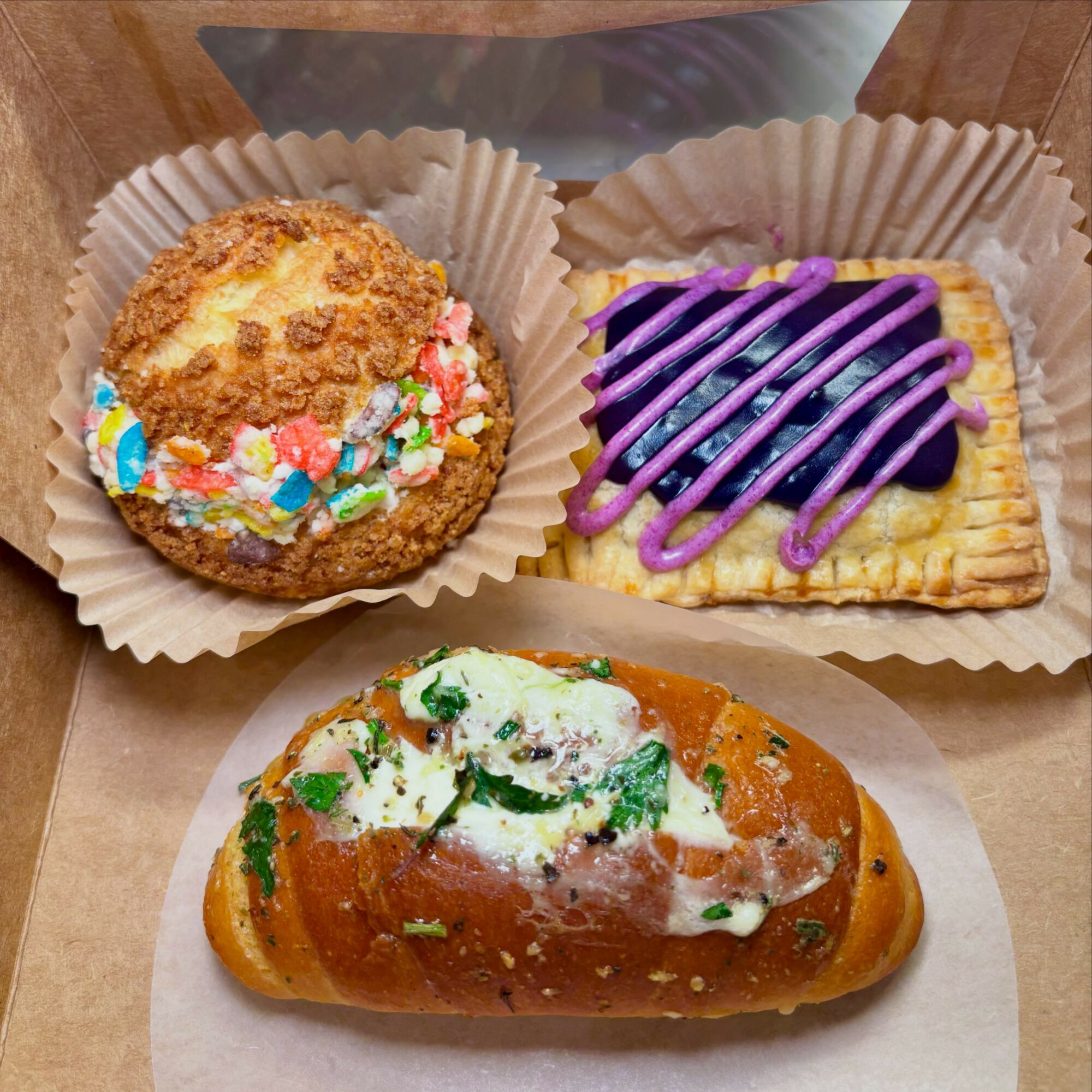 Two baked goods in paper cups and a pastry on a white paper napkin. One is a cookie with colorful sprinkles, the other is a cracker with purple icing and drizzle. The pastry is topped with white icing, green herbs, and small bits of vegetables.
