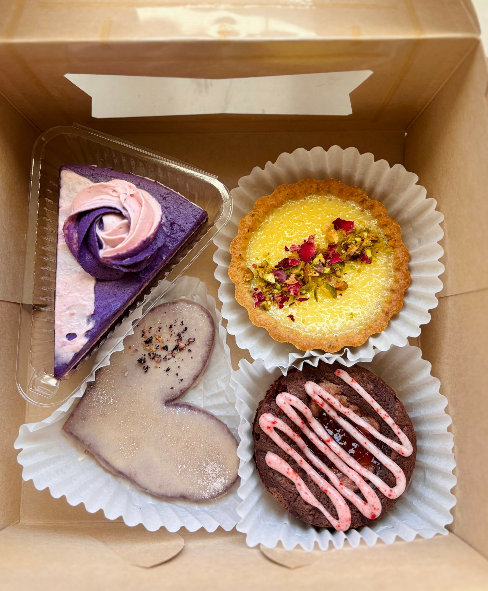 Four desserts in a box: a purple and white slice of cake, a yellow cupcake with red and green toppings, a heart-shaped cookie, and a chocolate donut with pink drizzle.