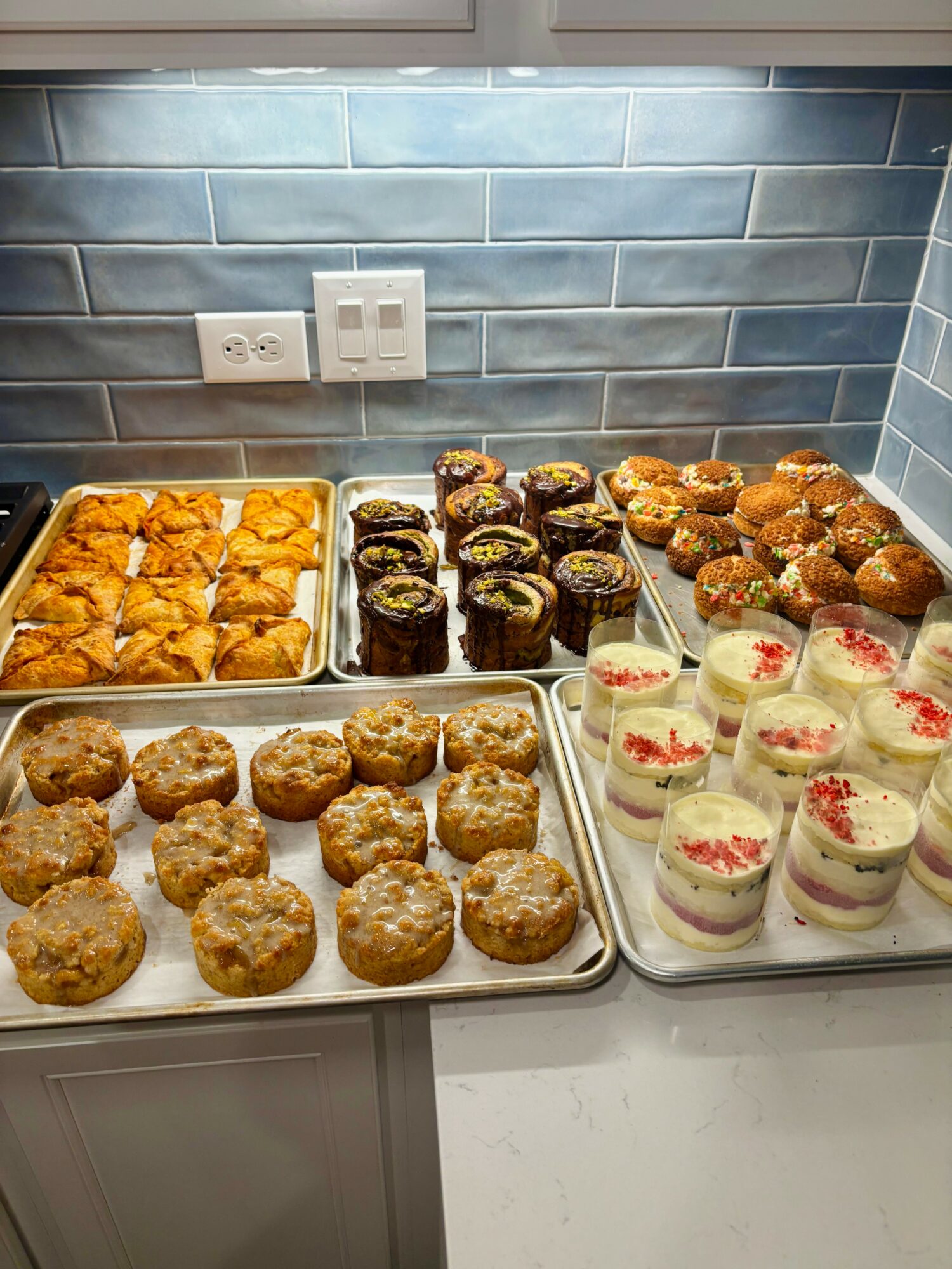 Tray of baked goods, including muffins, cupcakes, and pastries, on a kitchen counter with blue tiled backsplash.