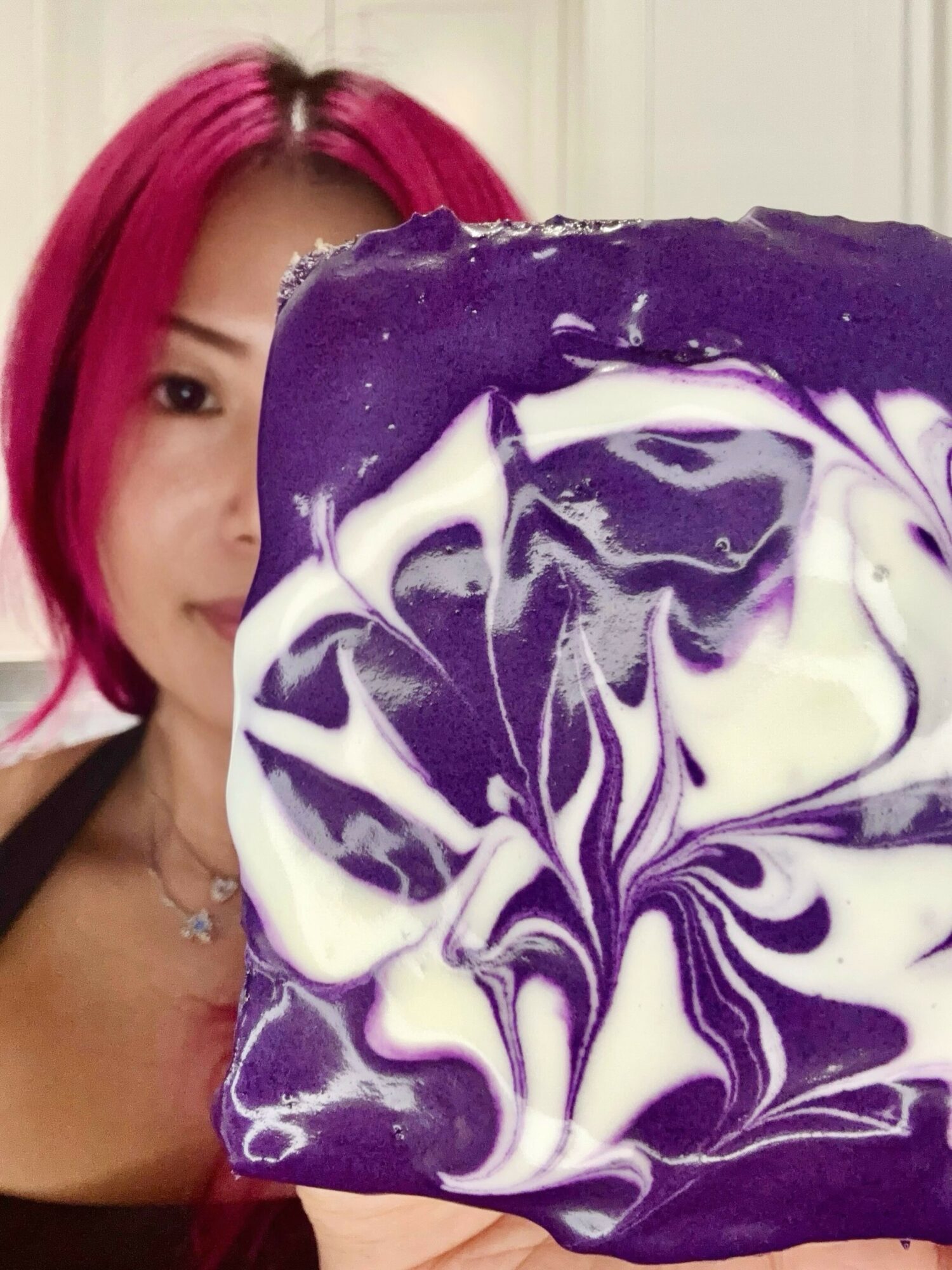 Person with pink hair holding a purple and white marbled soap or object, partially covering their face, in a room with white walls.