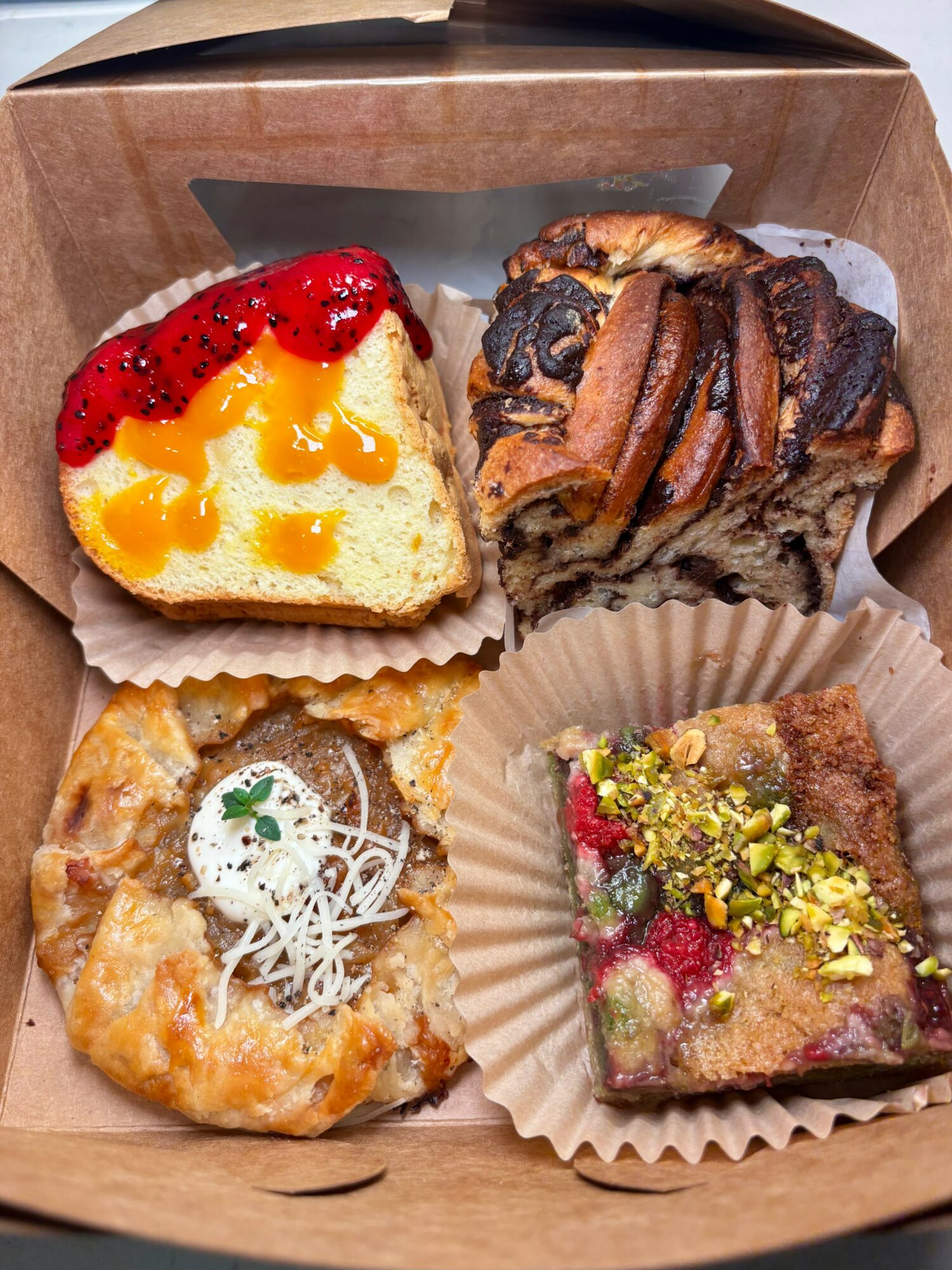 Four assorted baked goods in a box: a cheesecake with red fruit topping, a chocolate swirl pastry, a fruit-topped cake, and a pastry with nuts and fruit pieces.